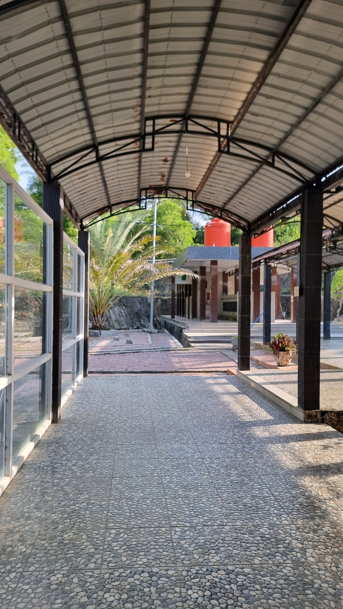 The image depicts a covered walkway with a corrugated metal roof supported by dark columns. The walkway floor is paved with small, grey, rounded stones laid in a grid pattern. To the left, a glass wall with white frames reflects the surrounding environment, including trees and the sky.
In the background, through the open end of the walkway, a building with multiple columns and a dark roof is visible. Two bright orange water tanks are situated on top of this building. The area beyond the walkway appears to be an outdoor space with paved pathways and some vegetation, including a palm tree. The scene is bathed in sunlight, casting shadows on the ground.
This location is identified as Blangrambong, Indonesia.