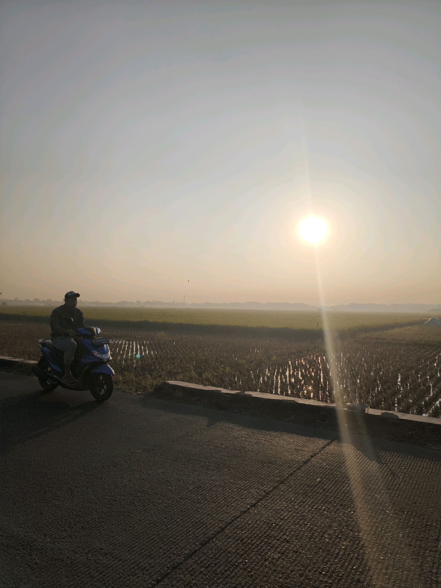 An individual wearing a dark long-sleeved shirt, light-colored pants, and a baseball cap is seated on a blue scooter. The scooter is positioned on an asphalt road adjacent to a rice paddy field. The road surface has a ribbed texture. A concrete barrier separates the road from the field, which contains shallow water and rows of harvested rice stalks. In the background, a vast expanse of green rice fields stretches towards a hazy horizon, with a faint silhouette of trees and structures. The sun is high in the pale sky, casting a bright glare and a noticeable sunbeam that extends downwards. The overall atmosphere suggests an early morning or late afternoon setting in Ciparay, Indonesia, characterized by natural light and rural scenery.