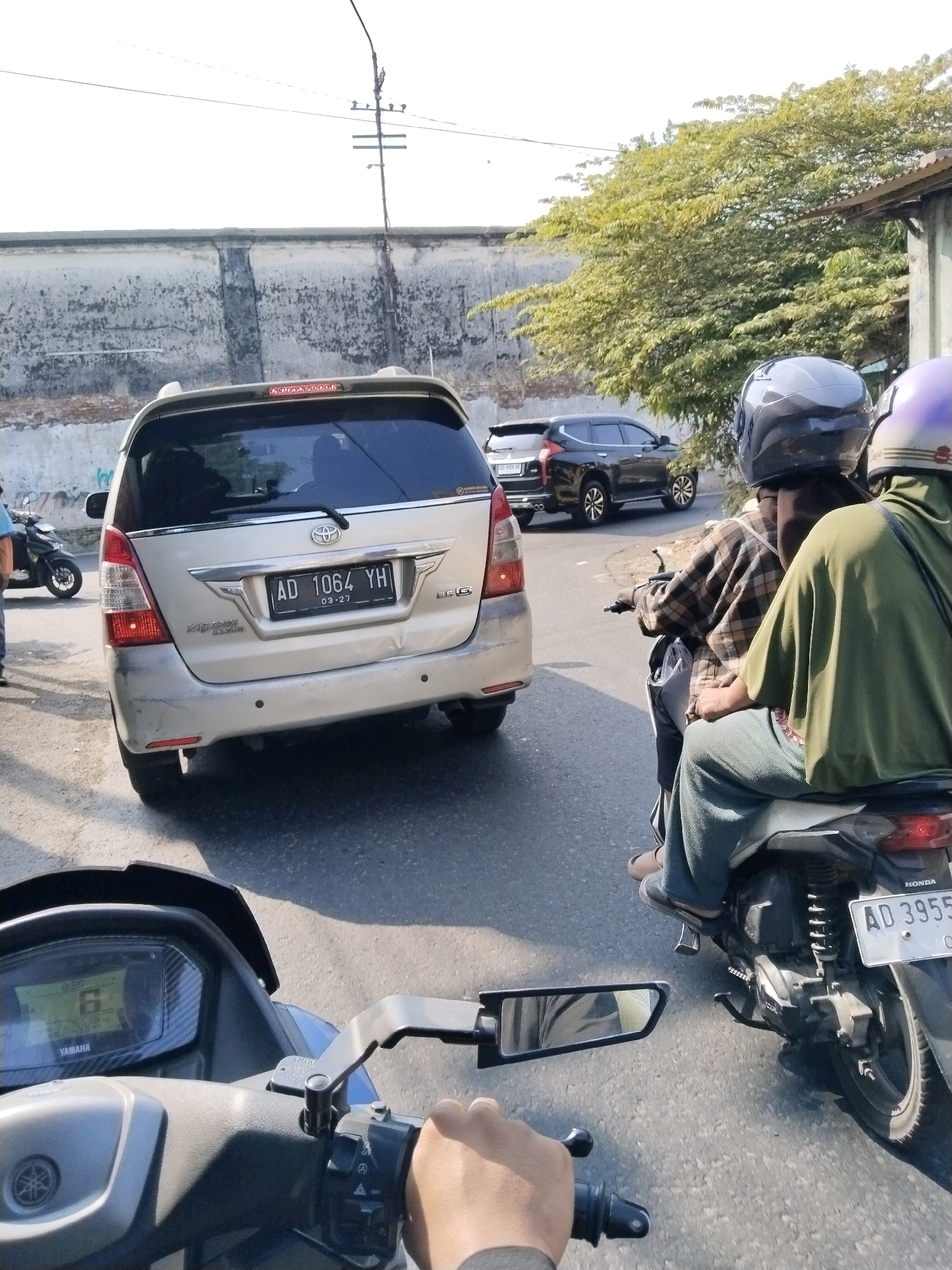 A sunny daytime view from the perspective of a motorcycle rider in Surakarta, Indonesia, captures a traffic scene. In the foreground, the dashboard of the motorcycle displays "6" in a large digital readout, along with "16528" and "YAMAHA" branding. To the right, the rider's hand is visible gripping the handlebar.
Further ahead, a silver Toyota Innova SUV with license plate "AD 1064 YH" is stopped in traffic. Behind the Innova, a dark SUV is also visible. Two individuals are riding a scooter in the lane to the right of the Innova. The rider in the front wears a plaid shirt and a dark helmet, while the passenger behind wears a green outer garment and a purple helmet. Their scooter displays license plate "AD 3955".
The environment includes a concrete wall on the left, partially covered with graffiti. A large tree with green foliage overhangs the scene on the right, near a simple building with a corrugated metal roof. Utility poles and wires are present in the background, suggesting an urban setting. The road surface is asphalt. The overall impression is a snapshot of everyday traffic in a city.