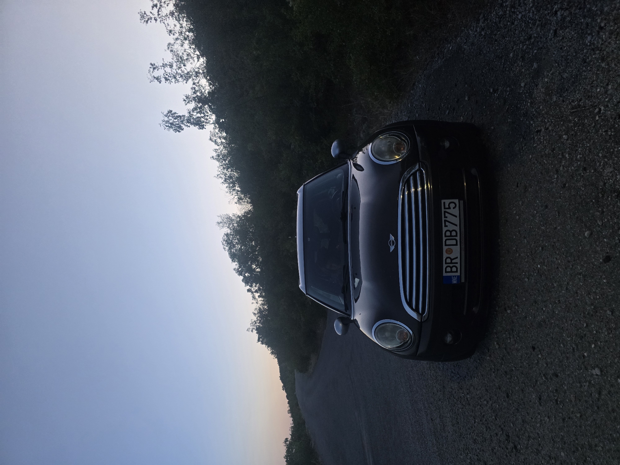 The image depicts a black Mini Cooper parked on a gravel shoulder next to an asphalt road. The car's front end is facing the camera. A license plate is visible, reading "BR DB 775" with "MNE" on the left side, indicating it is registered in Montenegro. The car is positioned on the right side of the road, which curves away into the distance to the left. The surroundings are characterized by dense vegetation on the right and a slightly hilly landscape on the left. The sky is clear with a gradient of blue and a hint of orange near the horizon, suggesting it is either sunrise or sunset. The overall lighting indicates it's a twilight scene. There are no people visible in the image. The location is identified as Klopot, Montenegro.