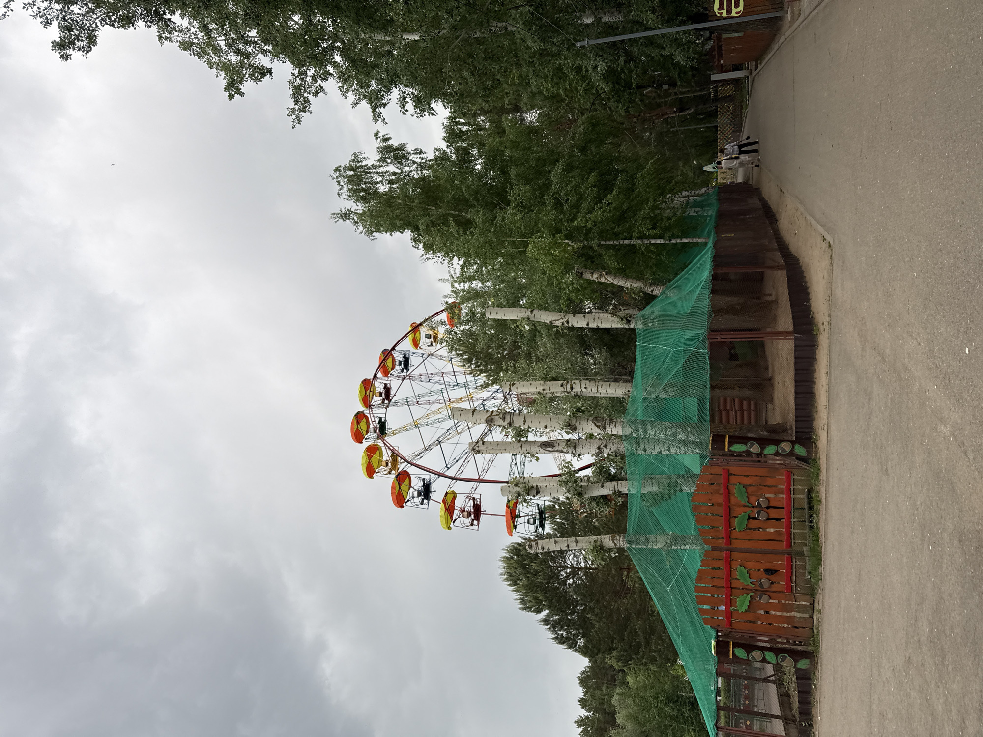 The image shows a Ferris wheel located in the city of Chaikovsky, Russia. The Ferris wheel is surrounded by birch trees and a green mesh fence. A paved walkway is visible on the right side of the image, with two people walking in the distance. The sky is overcast. A decorative wooden fence with leaf-shaped cutouts is present near the Ferris wheel.