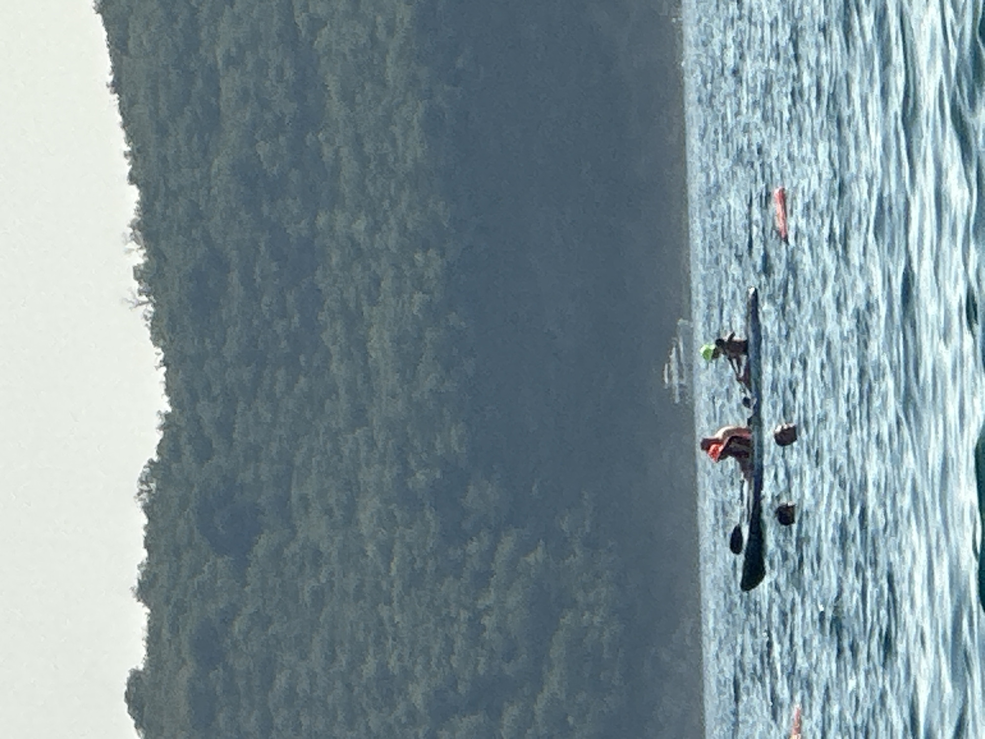 In Boreti, Montenegro, a serene body of water is dotted with individuals engaged in aquatic activities. The foreground of the image showcases the gentle ripples of the sea, with several people visible. Three heads emerge from the water, indicating they are swimming. A man in an orange life jacket is seated in a kayak, holding a paddle. Another person, wearing a green helmet, is also in the kayak. To the left, a person is partially visible with an orange flotation device. To the right, an orange flotation device floats independently on the water. In the middle ground, a small boat is anchored, its white hull contrasting with the deep blue of the water. The background is dominated by a lush, green hillside, densely covered with trees, forming a natural barrier that meets the hazy sky. The overall scene depicts a peaceful day at the beach with recreational activities.