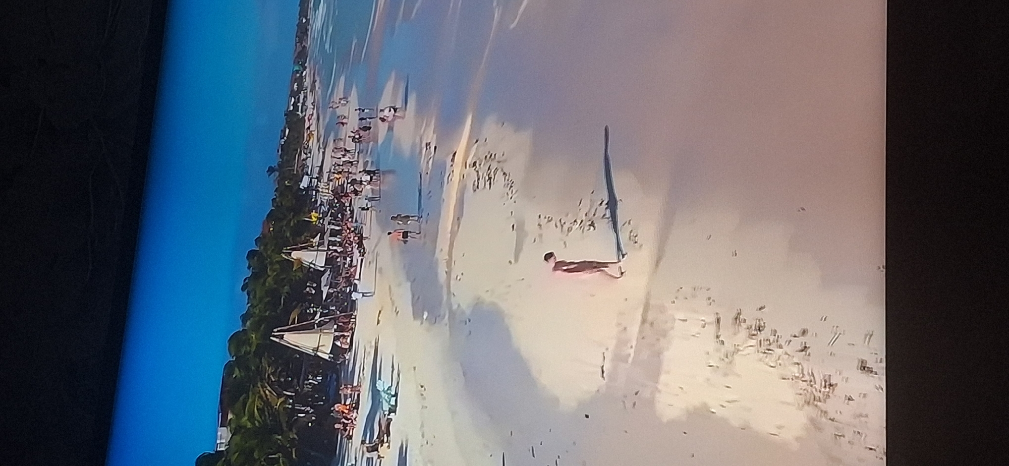 This is an aerial drone shot of a crowded beach with a body of water and a clear blue sky. The beach is lined with palm trees and several umbrellas and lounge chairs. There are many people scattered across the sand, some walking, some sitting, and some standing in groups. In the foreground, a single person is walking along the shoreline, casting a long shadow. The water is calm and a light blue color, with gentle waves breaking on the shore. The image does not depict any part of the city of Bila Tserkva, Ukraine.