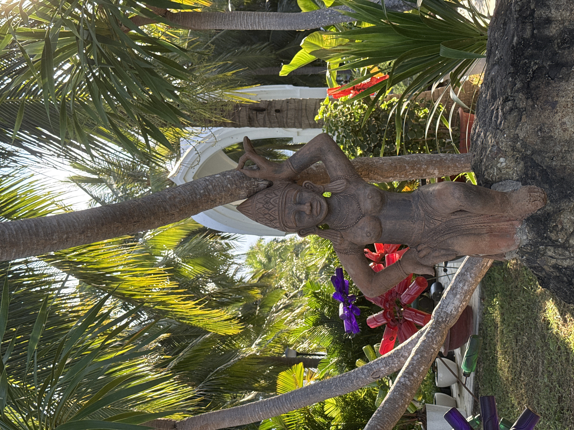 The image shows a statue of a woman in a garden in Nha Trang, Vietnam. The statue is made of stone and is wearing traditional clothing. The statue is sitting on a rock, and there are palm trees and other plants in the background. There is also a white structure in the background that looks like a gazebo or a temple. The sun is shining, and the lighting is bright, creating a warm and tropical atmosphere. The statue appears to be of Asian origin, possibly from Southeast Asia, given the context of Nha Trang. The overall scene suggests a tranquil, outdoor setting, likely a resort or a public garden, given the ornate statue and the lush greenery.