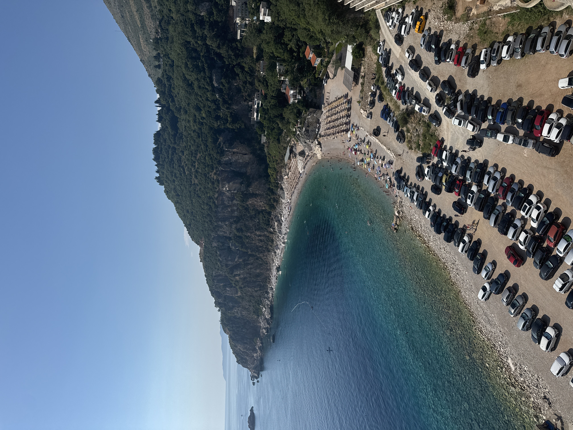 This is a high-angle, full shot of a crowded pebble beach and a large parking lot filled with vehicles, located in Mirište, Montenegro. The scene is bathed in bright sunlight, with a clear blue sky overhead. The turquoise waters of the Adriatic Sea gently lap against the shore, where numerous people are spread out on towels and under beach umbrellas. The beach itself is lined with rows of neatly arranged sunbeds.
The parking lot, situated adjacent to the beach, is densely packed with a variety of cars, predominantly SUVs and sedans, in different colors. Some people are visible walking between the parked cars and heading towards the beach. A steep, tree-covered mountain slopes down towards the sea, with buildings nestled amongst the greenery, indicating a coastal residential or resort area. The overall impression is of a popular tourist destination during peak season.