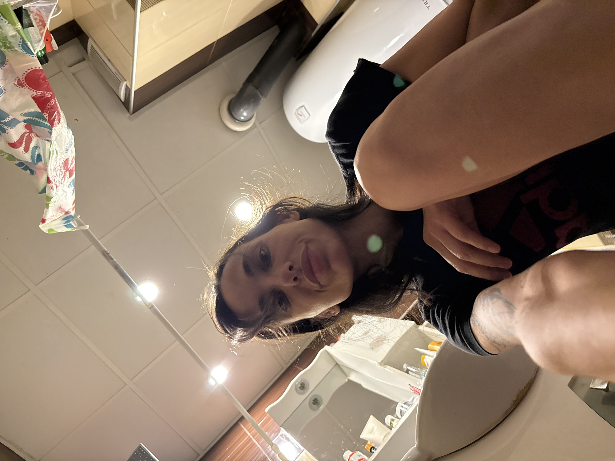 A woman is in a bathroom. She is holding a cell phone above her head and taking a photo looking down at herself. The woman has dark hair, light skin, and is wearing a black t-shirt. The bathroom is white and beige, with a tiled ceiling, sink, toilet, and a water heater on the wall. The location is Pleven, Bulgaria.