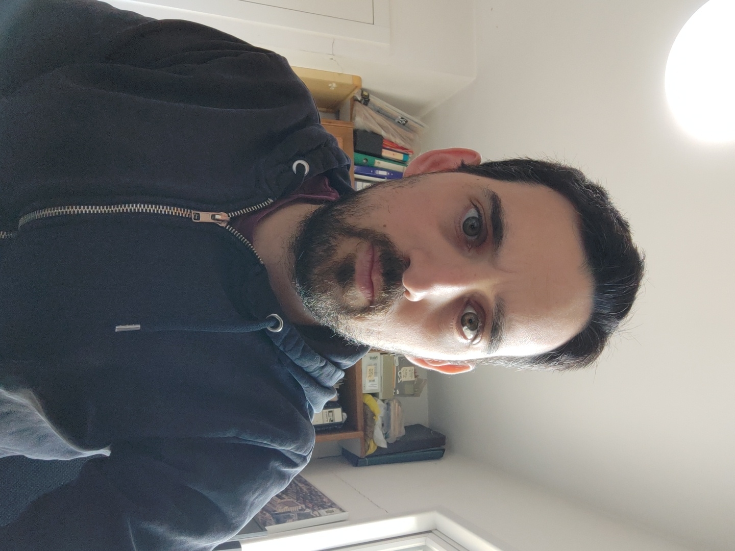 A male individual with dark hair, a short beard, and a mustache is depicted in a chest-up portrait, looking directly at the camera. He wears a dark blue zippered hooded sweatshirt over a maroon undershirt. The zipper is silver-toned, and the hood drawstrings feature silver tips. The scene is an indoor environment in Raanana, Israel, characterized by white walls and a white ceiling. An illuminated circular flush-mount light fixture is visible overhead. To the left, a segment of a light-colored wooden bookshelf contains various binders and objects. To the right, a white-framed window admits natural light, and a framed image is mounted on the wall.