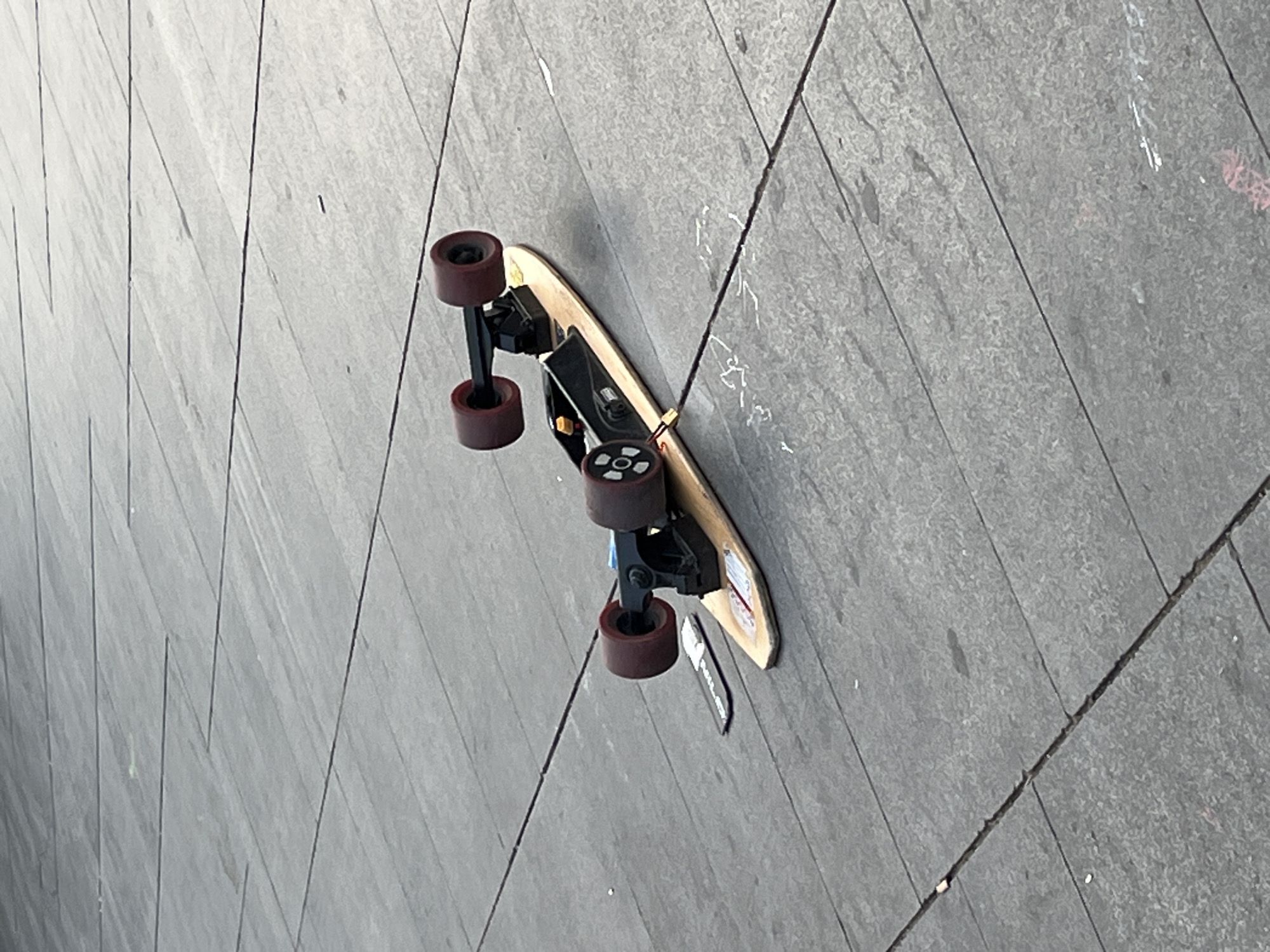 A longboard-style skateboard lies on its side on a grey paved surface, likely outdoors in Barcelona, Spain. The ground is composed of rectangular tiles or slabs arranged in a diagonal pattern.
The skateboard features a light wooden deck, dark red or maroon wheels, and black trucks. An electronic component, resembling a motor and battery pack, is mounted on the underside of the board between the trucks, indicating it is an electric skateboard. A separate black rectangular object, possibly a remote control, rests on the pavement next to the board's front wheels.
On the underside of the wooden deck, near the tail, a graphic or sticker is partially visible. It contains red elements and a white box with indistinct text that might include "E-VO," and potentially parts of words like "RACE" or "BOARD." The black object next to the board also displays blurred white text, which is difficult to decipher but might contain words like "PULL & GO."
The lighting suggests it is daytime, and the paved surface appears dry. The skateboard is stationary, with no discernible activity or people present in the frame. The scene presents an urban setting, possibly a sidewalk or plaza, with the board seemingly at rest.