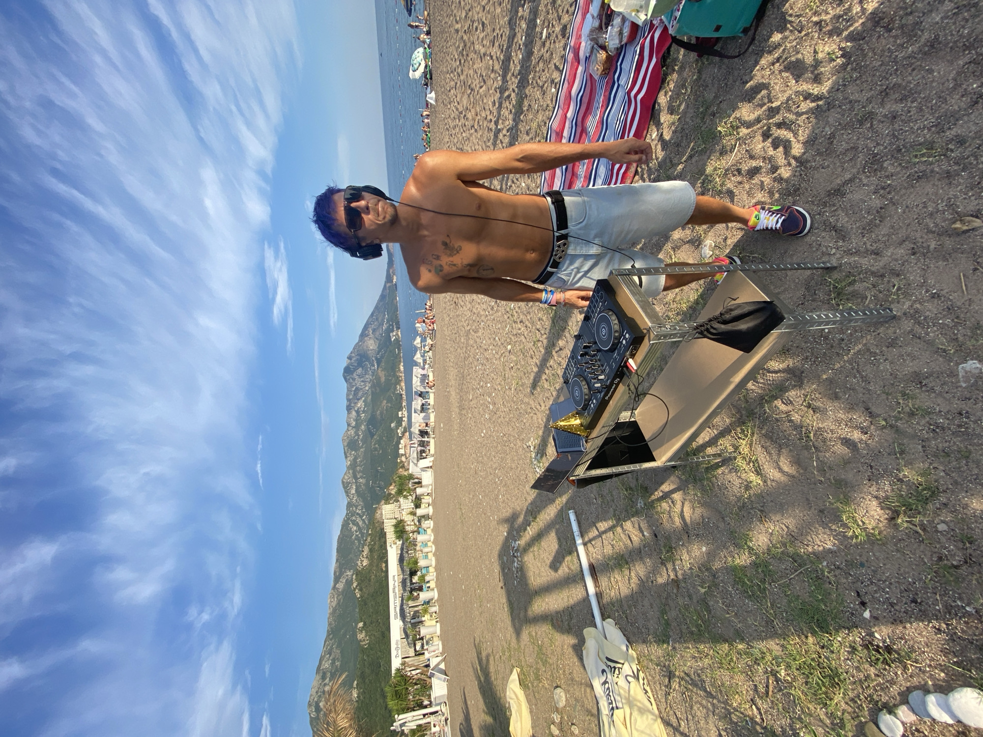A shirtless man with blue hair, sunglasses, and headphones stands on a beach, posing for a photo. He is standing next to a DJ setup that includes a mixer, laptop, and a black bag. The man is wearing light-colored shorts and colorful sneakers. In the background, there are lounge chairs, umbrellas, and people on the beach, with a body of water and mountains in the distance. The sky is blue with some wispy clouds. A red, white, and blue striped blanket is visible to the right of the man, along with a green bag. The scene appears to be set during the daytime, likely late afternoon, given the shadows. The man's pose and headphones suggest he is a DJ preparing to play or performing. There is no visible text in the image.