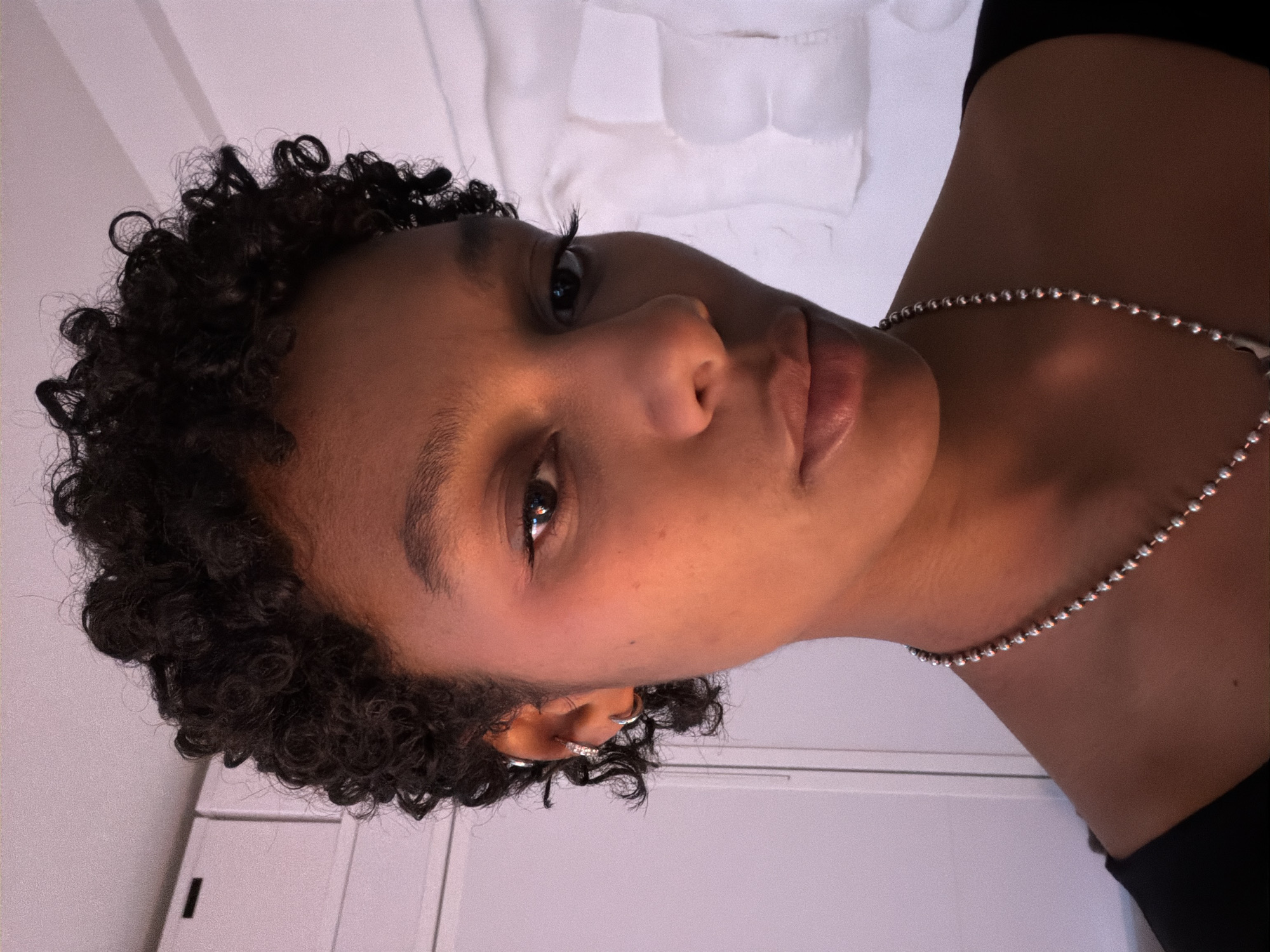 A person with short, curly, dark hair is seen at an oblique angle, facing the camera. They have light-brown skin, and their lips are full. A silver beaded necklace is visible around their neck. Their gaze is direct. The background includes a white surface, possibly a bed or other fabric, and a white door or wall. The location is not discernible as Paris, France.