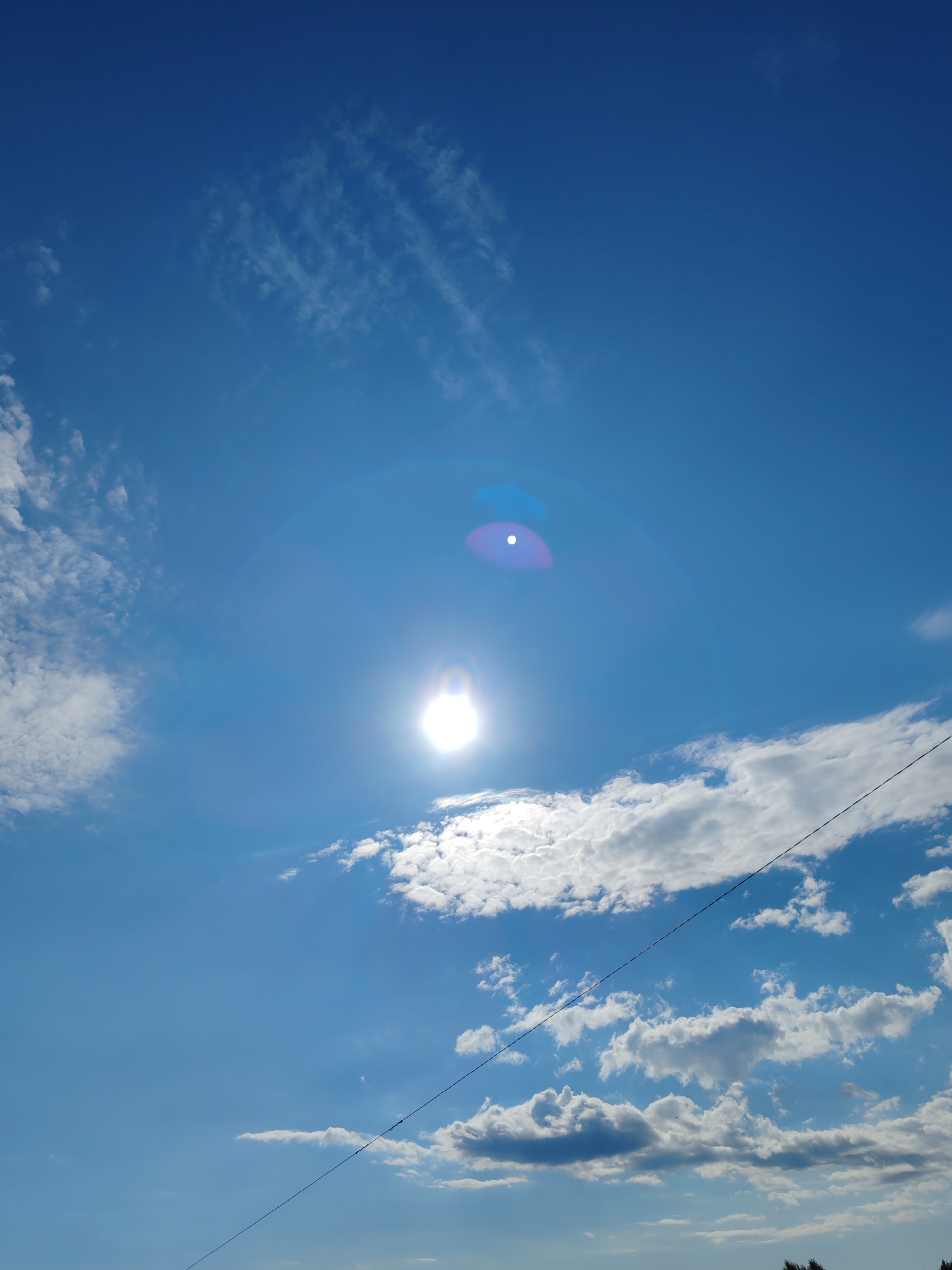 The image is a vertical, outdoor, daytime photograph looking up at a bright blue sky with scattered white clouds. The sun is visible as a very bright, blown-out light source with a lens flare creating concentric rings of light and color, predominantly purple and white. Wispy, cirrus-like clouds are present in the upper left portion of the sky, while larger, cumulus clouds are scattered throughout the mid-ground and lower portions of the image. A thin, dark power line runs diagonally from the upper right to the lower left of the frame, bisecting the sky. No people or specific landmarks are visible. The image does not provide enough information to determine its location within the city of Чухлэм, Russia.