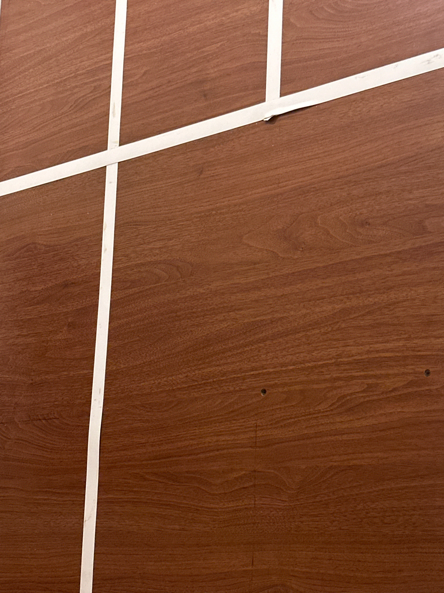 The image shows a close-up view of a wooden surface with white tape applied in a grid pattern. The wood grain is visible, indicating a brown, polished finish. There are two small, dark circular indentations visible on the wood surface towards the right side of the image. The white tape is applied in straight lines, forming intersecting sections. One horizontal piece of tape is near the top of the frame, and another runs diagonally from the top left towards the bottom right. A long, vertical piece of tape extends from the top center of the frame down to the bottom. The overall impression is that of a surface that has been marked or sectioned off using tape. The location in Kathmandu Metropolitan City, Nepal, is not discernible from the image alone.