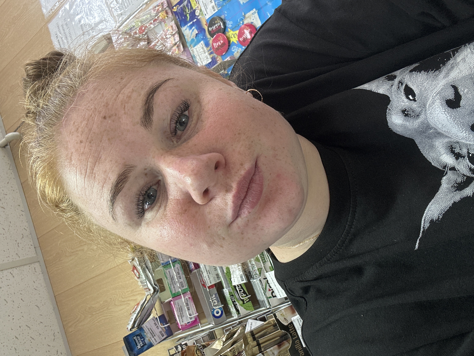The image is a close-up selfie of a young woman with light brown hair pulled up in a bun and freckles on her face. She is wearing a black t-shirt with a silver graphic of an animal on it and a delicate gold chain necklace. Her makeup includes mascara and lip gloss. In the background, there are shelves stocked with various products, including gum and coffee. The environment appears to be an indoor retail space, possibly a convenience store or supermarket, consistent with locations found in Chaikovsky, Russia, given the Cyrillic text visible on some of the product packaging. The lighting suggests an artificial indoor setting.