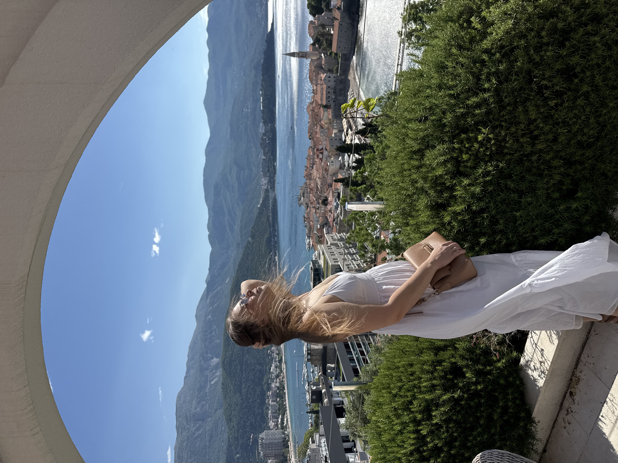 A vertical, outdoor, medium-shot photograph shows a woman wearing a white dress with a brown purse in the city of Budva, Montenegro. The woman stands outside with her left hand on her head and is looking to the sky in front of a greenery bush. The background features a coastline with mountains, water, and old buildings. The photograph is framed by a white arch in the upper left corner.