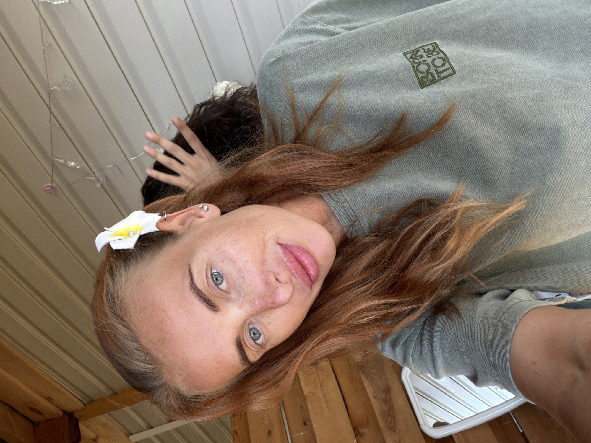 A woman with long, reddish-brown hair and blue eyes is taking a selfie. She is wearing a green t-shirt with the words "BORN TO BE" embroidered in green on the left chest. A white flower with a yellow center is tucked into her hair above her left ear. Her skin is lightly freckled. Behind her, another person's dark hair and hand are visible. The background appears to be an outdoor setting with wooden paneling and a white, corrugated ceiling. String lights are attached to the ceiling above and behind the woman. A white chair with striped upholstery is partially visible to the left of the frame. The lighting suggests it is daytime. The location is not identifiable as being in any specific city in Russia.
