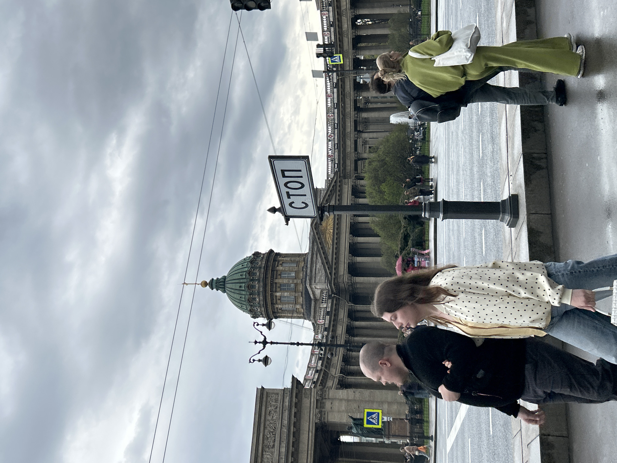 A medium shot of a street scene in Saint Petersburg, Russia, features the Kazan Cathedral in the background, with its distinctive green dome. A "СТОП" (STOP) sign is visible on a post in the foreground. Two people are walking on the sidewalk to the right of the frame. One person is wearing a black jacket and dark pants, and the other is wearing a white shirt with black polka dots and blue jeans. A couple is seen in the background on the sidewalk and street. The weather appears overcast, with a cloudy sky above. The buildings feature classical architectural elements, including columns and ornate facades. Wires are strung across the sky, and traffic lights are visible.