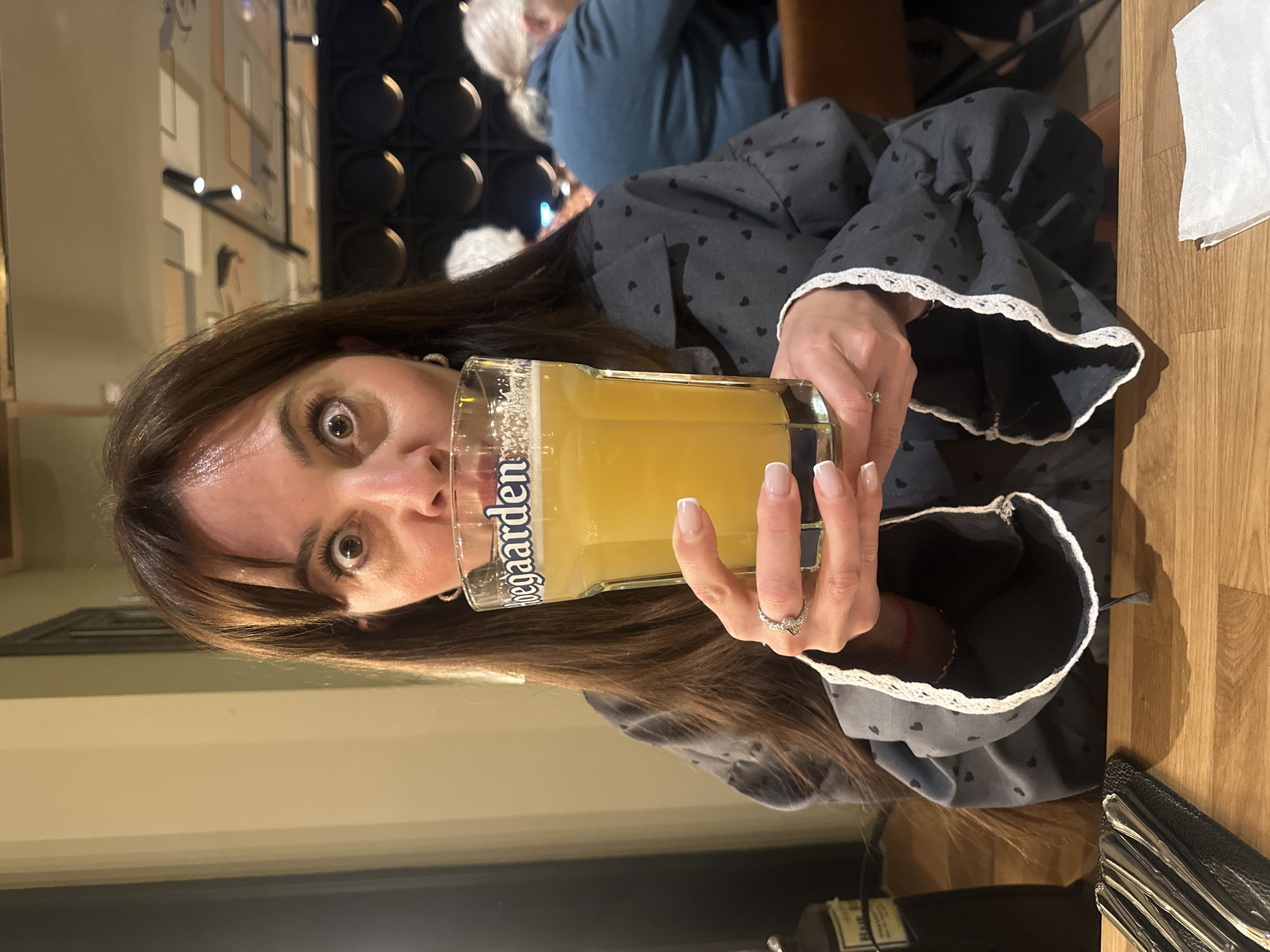 A woman is holding a glass of beer in front of her face. The glass has the word "Hoegaarden" on it. She is wearing a gray blouse with small black hearts and white lace trim. Her fingernails are manicured with a French tip. She is wearing a ring on her right ring finger. The background shows a blurred restaurant interior with black and white wall decorations. There is another person visible in the background. The location is Riga, Latvia.