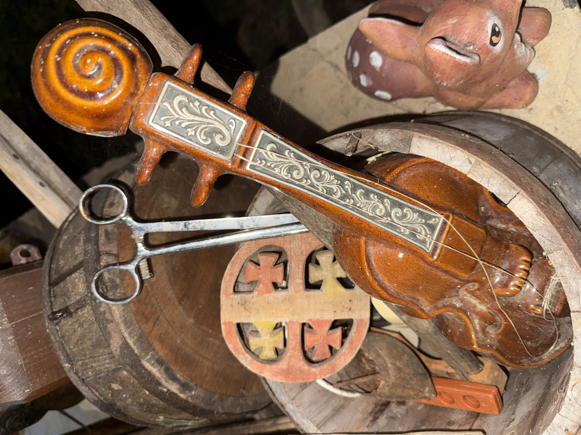 The image depicts a still life arrangement of various objects, including a ceramic violin, a wooden barrel, and a pair of metal scissors, situated in an outdoor setting that may be associated with Petrevene, Bulgaria, given the provided context. The violin, predominantly brown and orange in color, is placed within a cracked wooden bowl or trough. It features ornate detailing on its neck and scroll. The scissors, positioned above and to the left of the violin, appear to be surgical or crafting implements. A circular wooden object adorned with cross shapes in red and yellow is visible behind the violin. To the right of the bowl, a small, reddish-brown ceramic figurine of a fox-like creature with white spots is present. The background consists of wooden structures and appears to be an outdoor environment, possibly a yard or workshop. The lighting suggests it is either dusk or nighttime. There are no people or animals actively interacting with the objects.