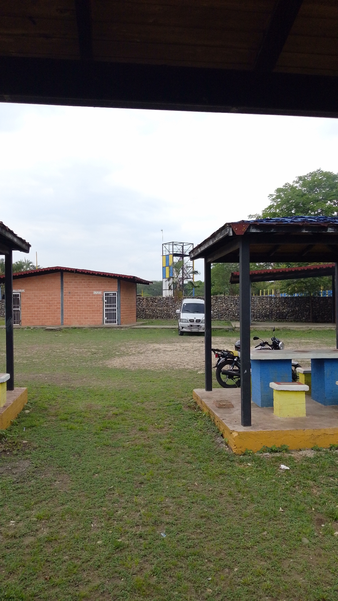 The image shows an outdoor recreational area in Tinaquillo, Venezuela. In the background, there is a light-colored van parked near a stone wall and a metal tower structure painted in blue, yellow, and black. There are two brick buildings with metal doors, labeled "HILEROS" and "DAMAS". A motorcycle is parked under a shaded pavilion with a concrete picnic table painted blue and yellow. The area is covered in grass. The sky is overcast. The photo is taken from under a partially visible wooden roof.