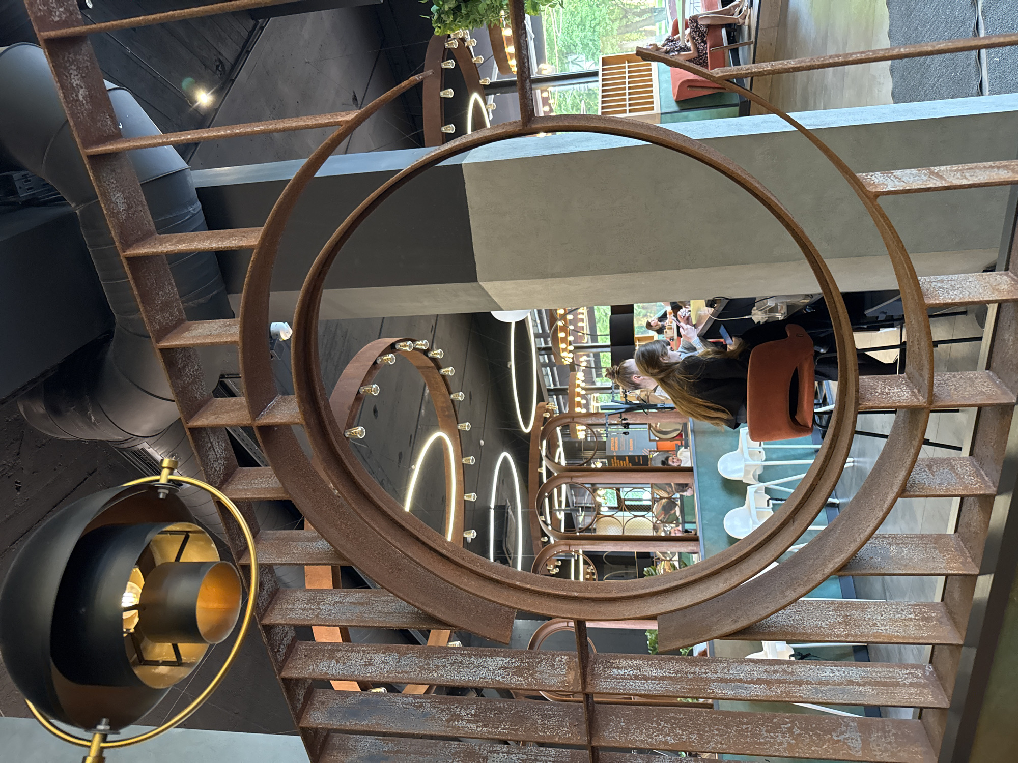 This image captures the interior of a contemporary cafe, likely located in Veliky Novgorod, Russia, given the provided context. The foreground is dominated by a large, circular, rust-colored metal structure, likely a decorative element of the cafe's architecture. Through this structure, a glimpse of the cafe's interior is visible.
In the mid-ground, a woman with long brown hair, wearing a black top, is seated at a table, looking down at her phone. To her left, another person, also with brown hair, is visible, seemingly engaged with their phone. Several other patrons are visible in the background, some seated at tables and others standing. The cafe features modern lighting, including ring-shaped LED fixtures and exposed bulb pendant lights. The walls are a mix of dark grey and light grey concrete. Green accents are visible in the form of plants and a teal-colored wall. Several high chairs are also present.
The overall impression is of a modern, well-lit cafe designed with industrial and contemporary elements. The specific location in Veliky Novgorod, Russia, is inferred from the prompt's directive.