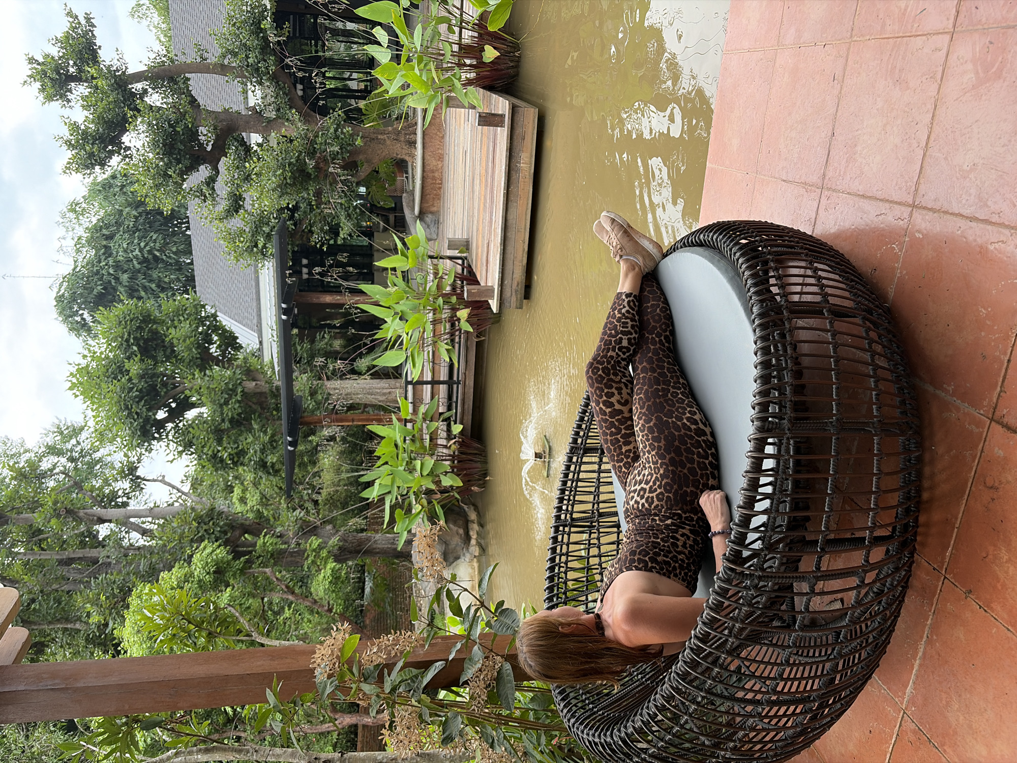A woman wearing a leopard print jumpsuit and tan sneakers is reclining on a large, round, dark wicker chair with a gray cushion. She is facing away from the camera, looking towards a pond with a small fountain in the center. The chair is situated on an orange tiled patio. Behind the patio is lush greenery, including trees and large leafy plants. In the background, there is a building with a dark roof and what appears to be a wooden deck extending over the pond. The location is identified as Mae Rim, Thailand.