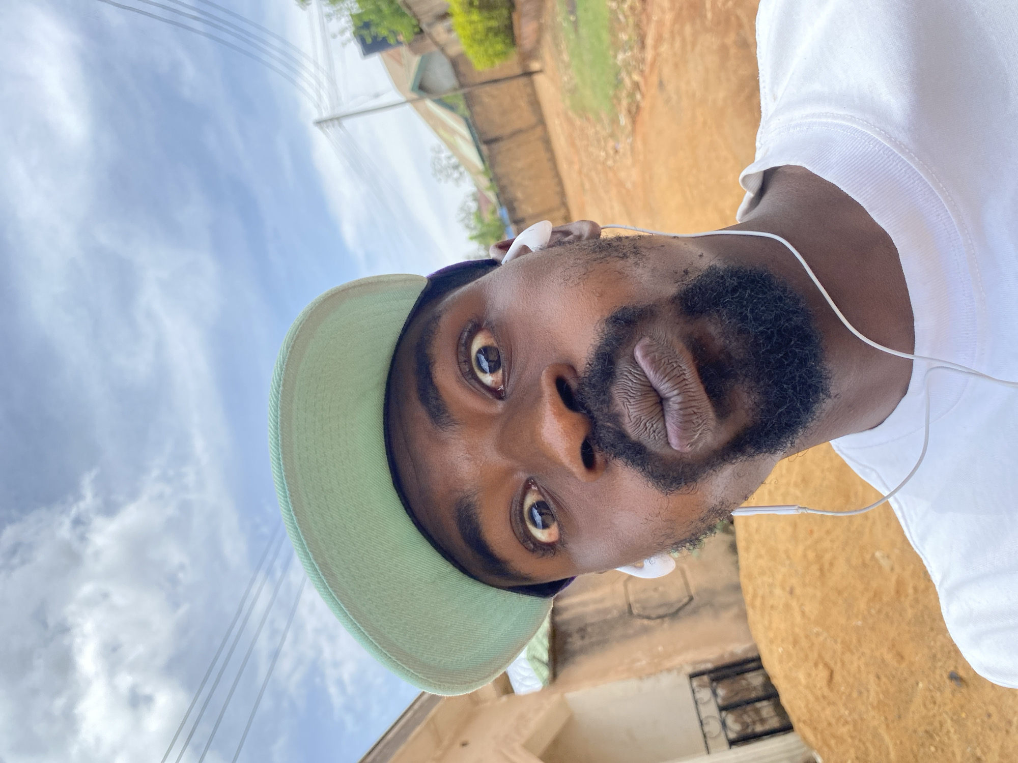 This portrait-oriented image features a close-up selfie of a young adult Black male, captured with a significant clockwise rotation. The man gazes directly into the camera with wide, expressive eyes, and his lips are playfully pursed. He sports a full beard and mustache. He is wearing a light-colored (likely white) t-shirt or sweatshirt and a light green baseball cap positioned backward on his head. White wired earphones are clearly visible in his ears, with the cable draped across his shoulder and chest.
The background reveals an outdoor setting under a bright, partly cloudy sky, indicating daytime. Multiple thin utility lines traverse the sky. Below, the ground transitions to a light brown, unpaved or dusty surface. Patches of green foliage and distant building structures are also discernible, suggesting a residential or semi-rural environment. The scene depicts a casual moment, possibly while walking or standing outdoors in Jos, Nigeria, with the man seemingly listening to audio through his earphones. There is no visible text in the image.