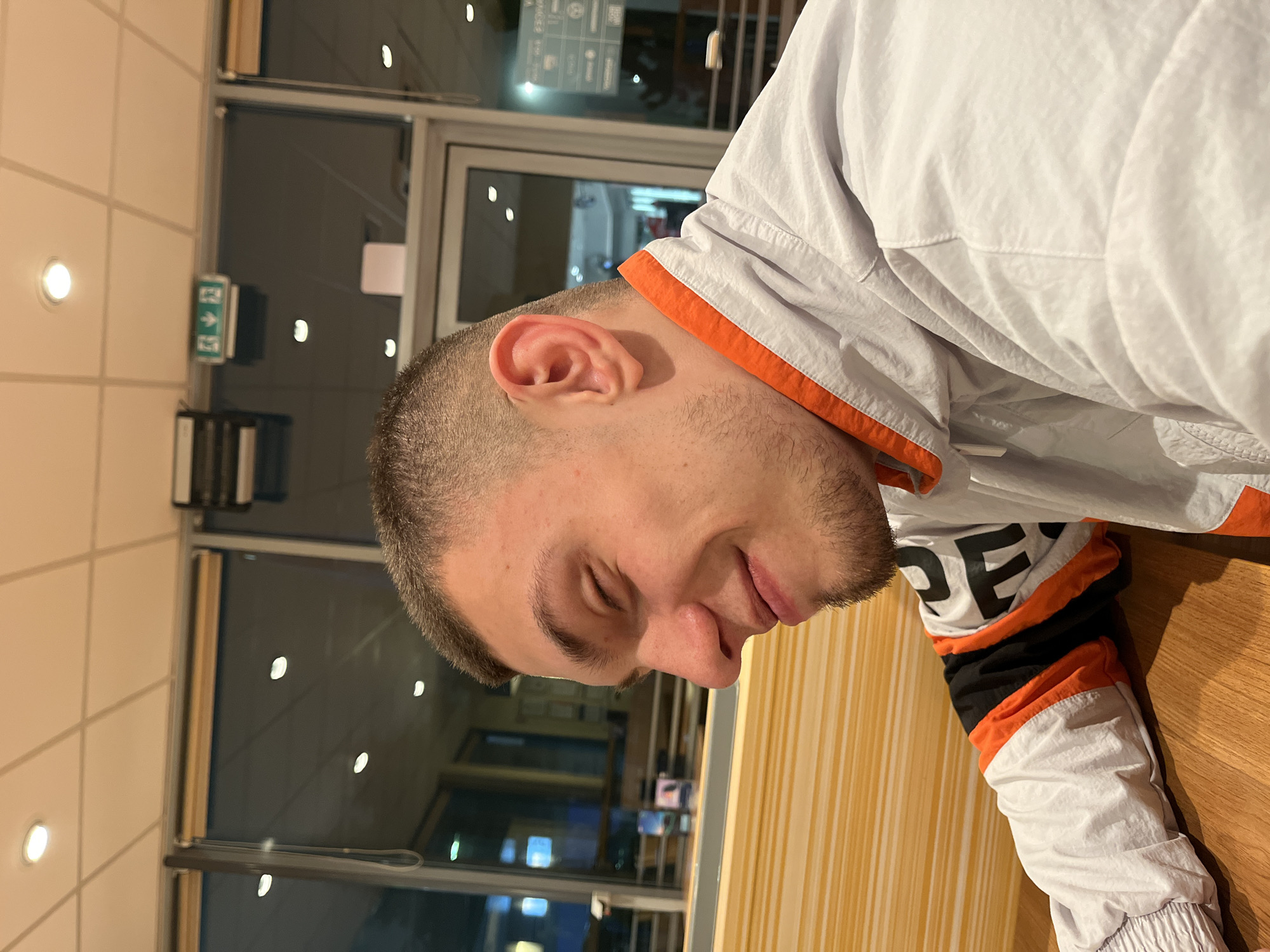 A man with short brown hair is seen in a close-up, angled shot. He appears to be indoors, with a wall and a window visible in the background. The window reflects lights that suggest an urban environment. He is wearing a light-colored jacket with orange and black accents, possibly a tracksuit top. The setting seems to be a cafe or a restaurant because of the wooden table. There's no concrete evidence the location is Burgas, Bulgaria.