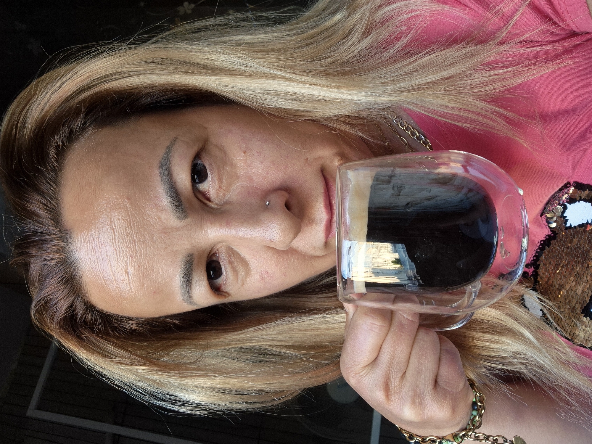 A medium shot shows a person holding a clear glass mug of dark liquid up to their mouth. The person has light brown hair with blonde highlights, and is wearing a bright pink shirt adorned with sequins. They also wear a gold chain necklace and a bracelet with green and gold beads. The background is out of focus, appearing to be an indoor setting with a window and a dark wall. The lighting suggests it may be daytime.