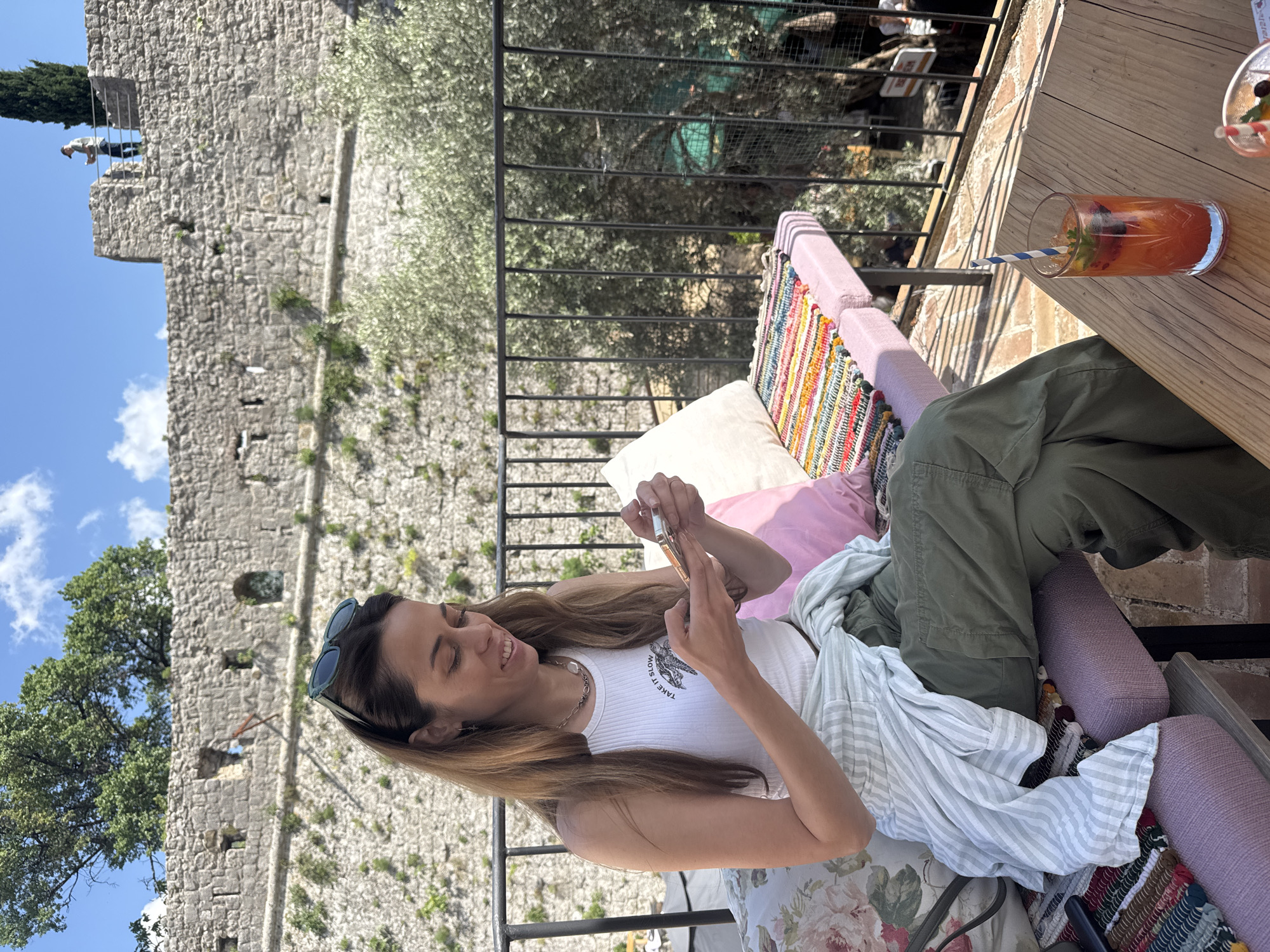 A medium, eye-level shot shows a woman sitting on a colorful cushioned seat at an outdoor table, looking at her phone. She wears sunglasses perched on her head, a white tank top, and olive green pants. A striped shirt is draped over her lap. Next to her is a wooden table with a glass of a reddish-orange beverage with a blue and white striped straw. The background is a stone wall on the left, and a metal railing overlooking trees and vegetation on the right. In the far left background on the top of the stone wall two human figures are standing on a parapet of the ancient fortress of Stari Bar, Montenegro.