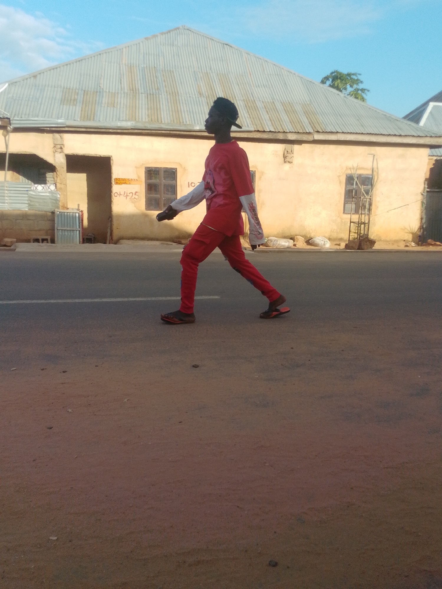 A young Black male wearing a red tracksuit and a black cap walks on the right side of a paved road in Gombe, Nigeria. The male is walking from left to right across the frame, with his left leg forward and his right leg extended behind him. He is wearing black and red sneakers. Behind him, a beige building with a corrugated metal roof is visible. The building has two windows and a doorway. There is a white line painted on the road in front of the building. The sky is a clear blue. The ground in the foreground is reddish-brown dirt.