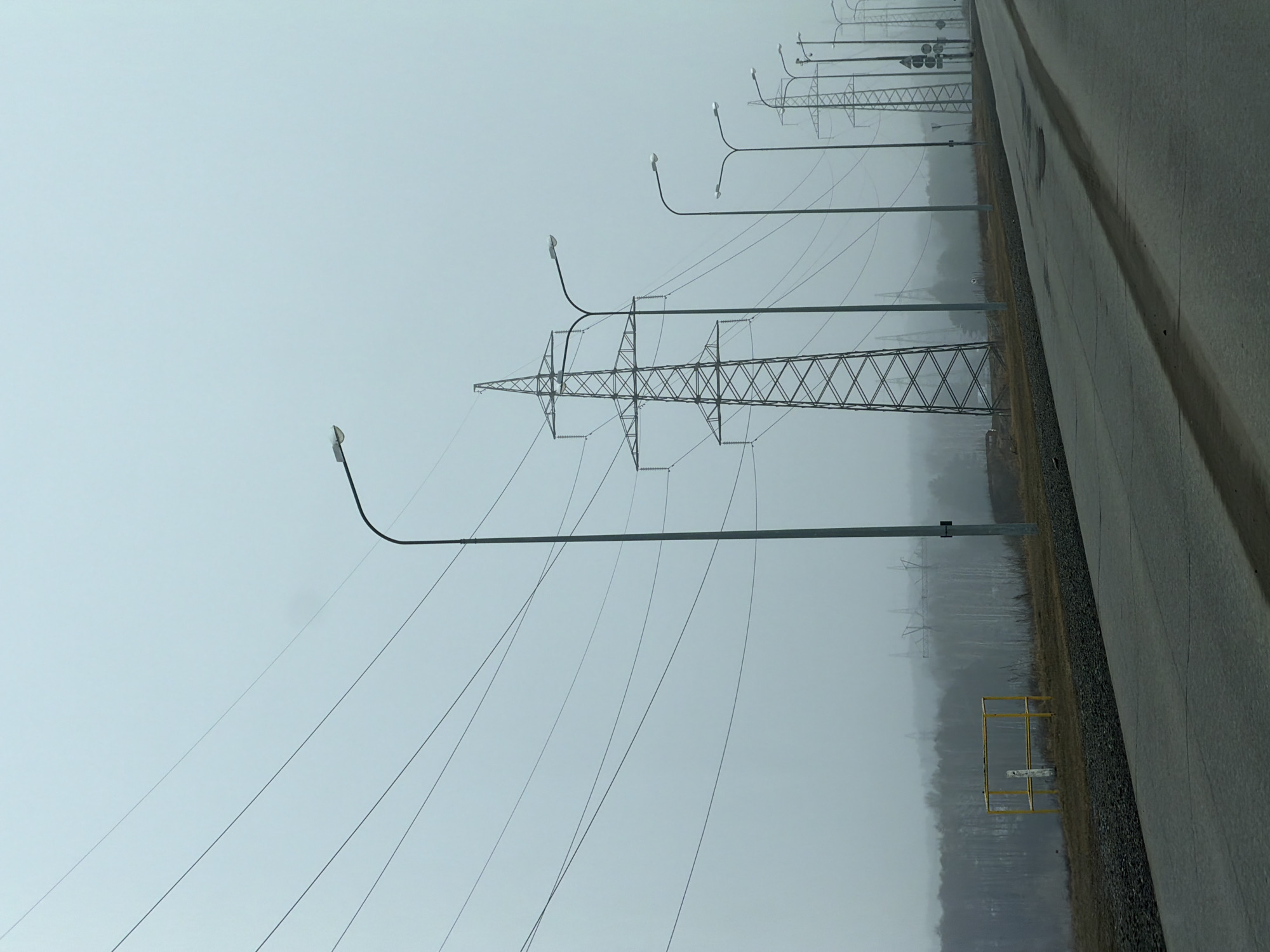 The image depicts a street scene in the city of Chaikovsky, Russia, on a misty or foggy day. Along the right side of the image is a paved road. The left side of the road borders a field with utility poles and power lines running along the landscape. Streetlights are spaced intermittently alongside the road and power lines. In the background, trees are visible through the mist or fog. A yellow metal structure is partially visible near the edge of the road.