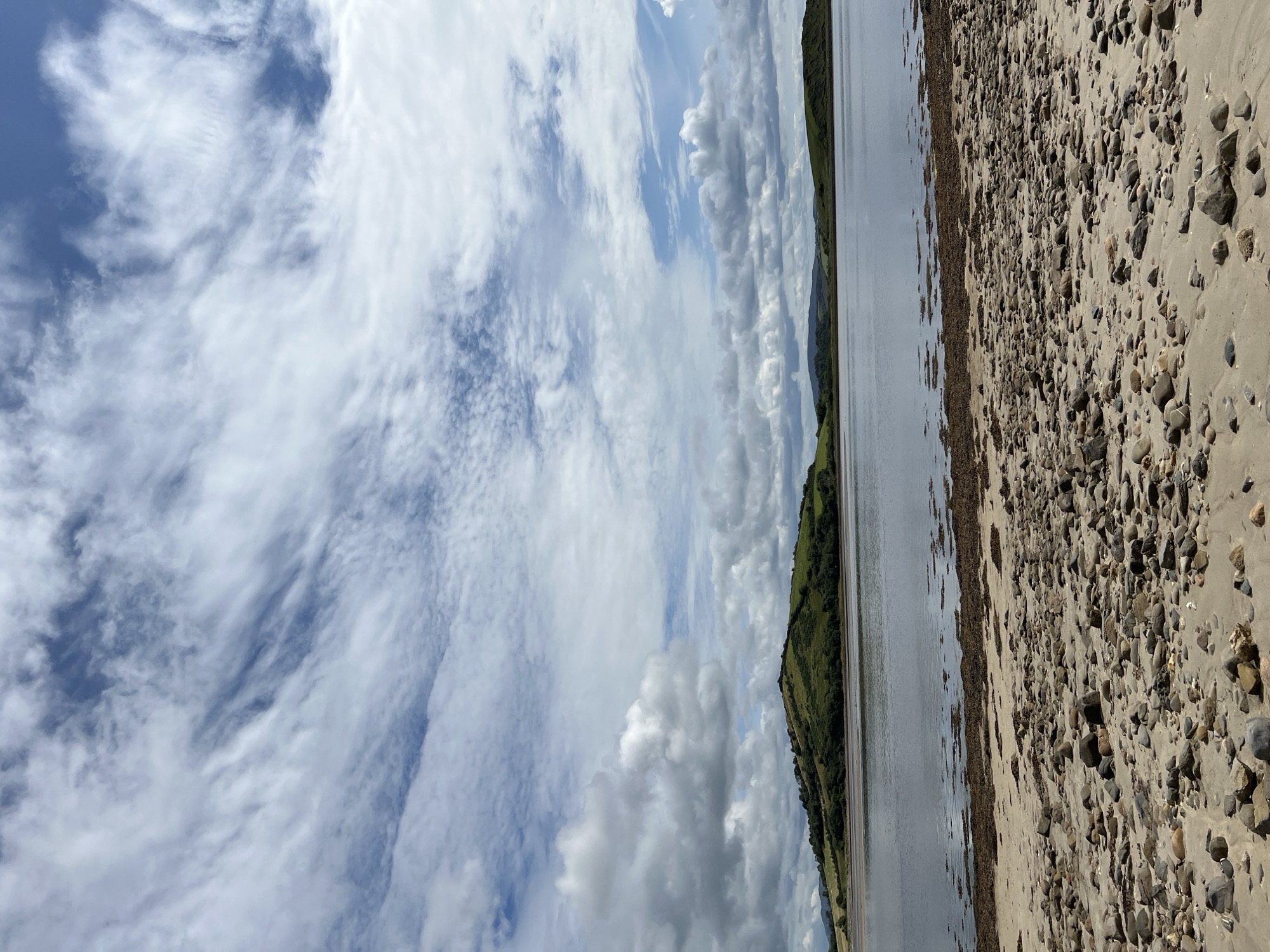 A wide-angle shot captures a serene coastal landscape under a dramatic, cloudy sky. The foreground is dominated by a sandy beach strewn with numerous smooth, grey and brown pebbles of varying sizes. A strip of dark seaweed lies along the tideline. Beyond the beach, a calm, wide body of water reflects the sky, with scattered patches of darker seaweed visible beneath the surface. In the middle distance, a lush, green, rolling hill rises from the water's edge, dotted with darker vegetation. The background shows more distant, gently sloping hills, also covered in green. The sky is a dynamic mix of bright white, wispy clouds and patches of deep blue, with clouds forming streaks and larger formations. No people or man-made structures are visible. The scene is likely Dunuisce, Ireland, a coastal town.