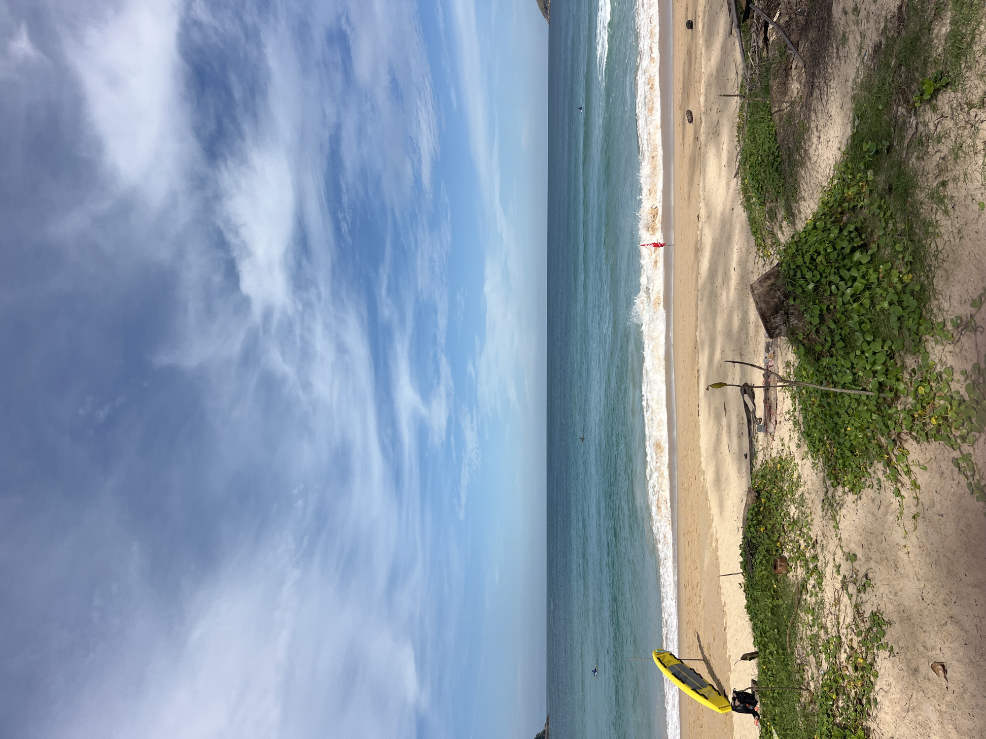 A wide-angle shot captures a serene beach scene in Rawai, Thailand, under a bright, partially cloudy sky. The turquoise ocean stretches to the horizon, with gentle waves lapping the sandy shore. A bright yellow surfboard stands upright on the beach near the water's edge, leaning against a dark backpack. A few surfers are visible in the distance, enjoying the waves. On the beach, a red flag stands as a marker. The foreground is dotted with lush green vegetation growing on sandy dunes, interspersed with driftwood and a tree stump. The overall ambiance is one of tranquility and natural beauty.