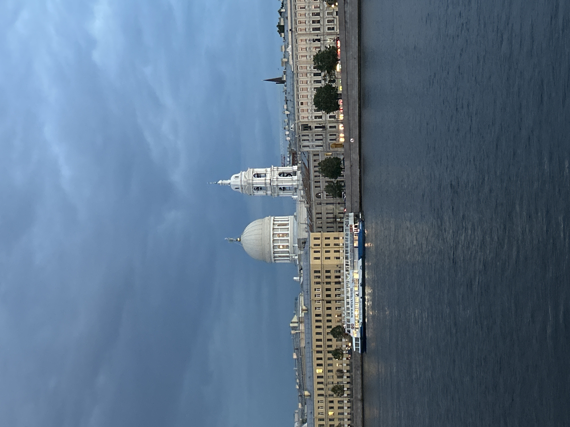 A panoramic, eye-level, full shot captures a wide river in the foreground, with its dark blue water reflecting the twilight sky. On the river's bank, a white and blue passenger boat named "WWW.NEVA.TRAVEL" is docked, its windows illuminated. Behind the boat, a long, neoclassical building with a pale yellow facade and numerous windows stretches across the scene. Further back, a prominent white dome with a golden finial crowns a large building, and to its right, a tall white bell tower with arched openings stands. The sky above is a dramatic expanse of dark, moody clouds. The overall scene suggests a late evening or dusk in a European city with significant architectural landmarks.