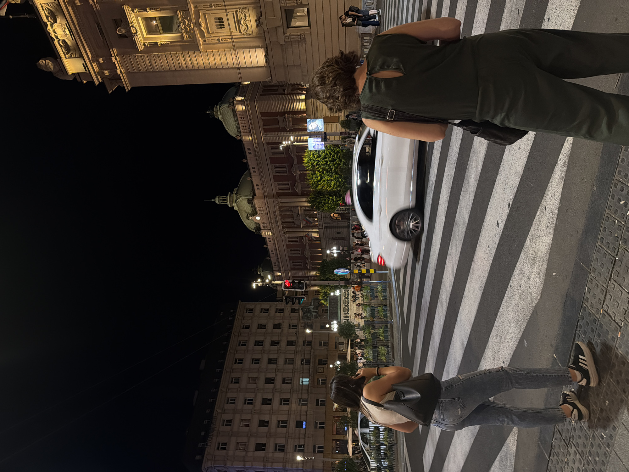 The image captures a nighttime street scene in Belgrade, Serbia, specifically in front of the National Museum. Two women are standing on a pedestrian crossing, waiting to cross the street. The woman on the left, facing away from the camera, wears a light-colored tank top and ripped blue jeans, carrying a black shoulder bag. She appears to be looking at her phone. The woman on the right, also facing away, is dressed in a dark green jumpsuit and carries a dark shoulder bag. A white car is captured in motion, crossing the pedestrian area. Behind the women, illuminated buildings with distinct architecture stand out, including the National Museum with its domes and the building to its right, featuring a large inscription that appears to be "1989". The street is lit by streetlights, and the overall atmosphere is that of a bustling urban environment at night.