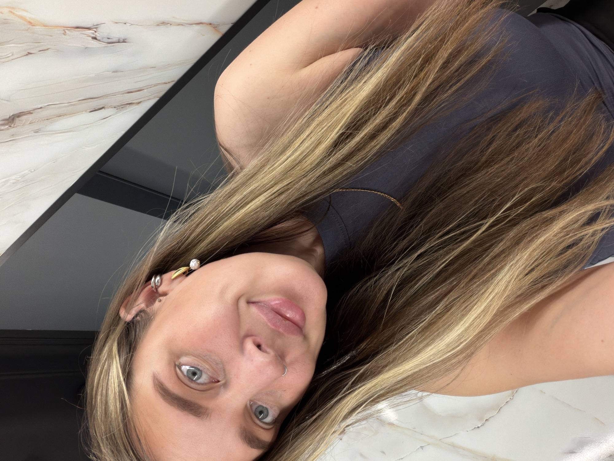 A woman is photographed in a selfie. She has long, highlighted blonde hair, light skin, and blue eyes. She is wearing a gray tank top and a small gold necklace. She also has several piercings in her left ear and a nose ring. The background consists of a white marble countertop and a black cabinet or wall. The location is not evident from the image. There is no indication that the photo was taken in Moscow, Russia.