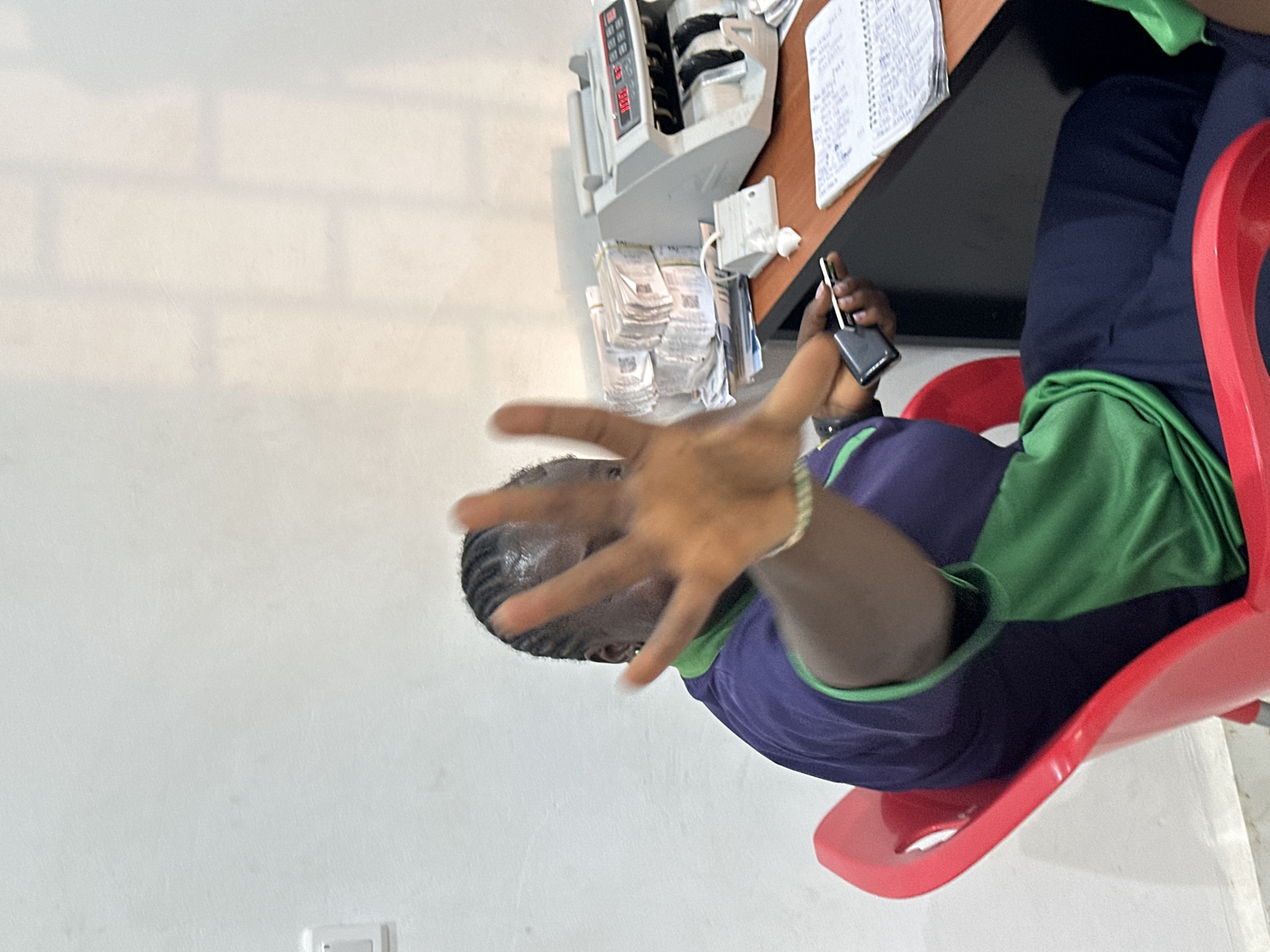 The image shows a person with dark skin sitting in a red plastic chair in what appears to be an office setting. The person is wearing a purple and green shirt and dark blue pants. Their right hand is extended toward the camera. In their left hand, they are holding a cell phone. To the left of the person is a wall painted white. A money counting machine and stacks of paper currency are visible on a desk next to the person, along with a notebook. The scene is in Bukuru, Nigeria.