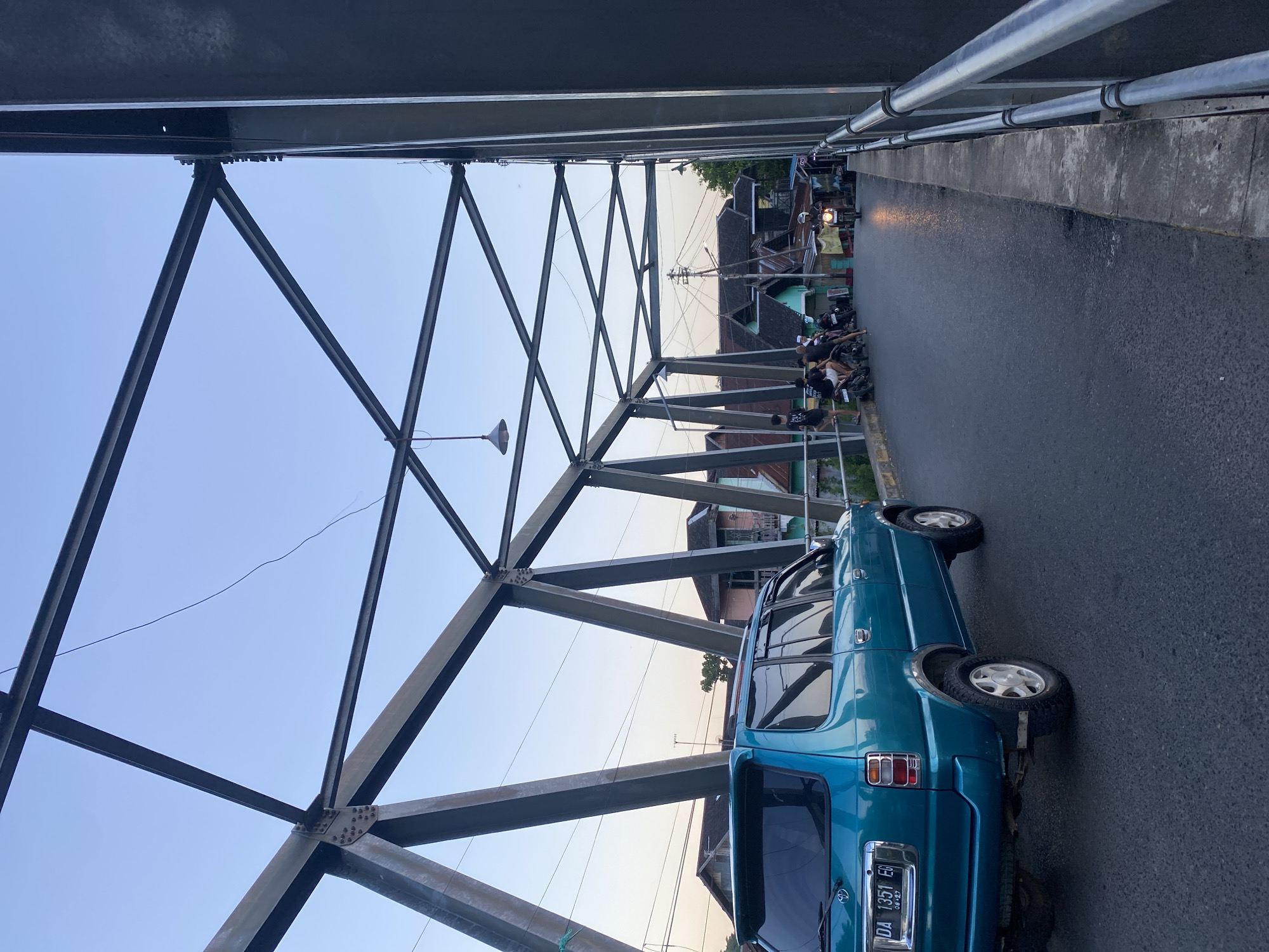 An outdoor, low-angle, eye-level shot captures a bridge's metallic truss structure against a clear, pale blue sky at dusk. On the bridge's asphalt roadway, a turquoise SUV is parked on the left, facing away from the camera. Its license plate reads "DA 1351 EG". In the background, several people on motorcycles are stopped on the side of the road, some standing and conversing, while others are seated on their bikes. A motorcycle is approaching in the distance, its headlight illuminated. The scene is consistent with the Tunggul Irang Ulu bridge in Indonesia, based on its architectural style and the presence of local vehicles and infrastructure.