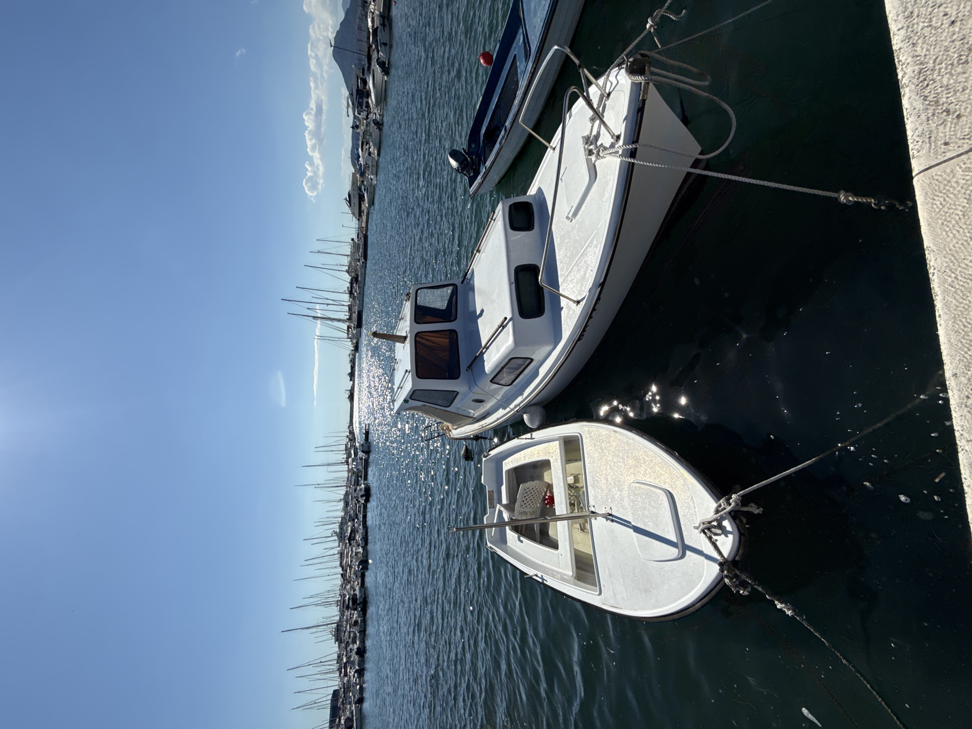In the city of Bar, Montenegro, on a clear sunny day, several boats are docked in a marina. In the foreground, two small white boats are tied to a dock. The boat on the left is smaller and has a single red seat inside. The boat on the right is larger and has a cabin. Behind these boats, a vast expanse of water is filled with numerous sailboats and motorboats, their masts creating a forest of vertical lines against the bright blue sky. The sun reflects off the water, creating a shimmering effect. In the distance, a mountainous landscape is visible under the hazy sky. The overall scene depicts a peaceful and picturesque harbor.