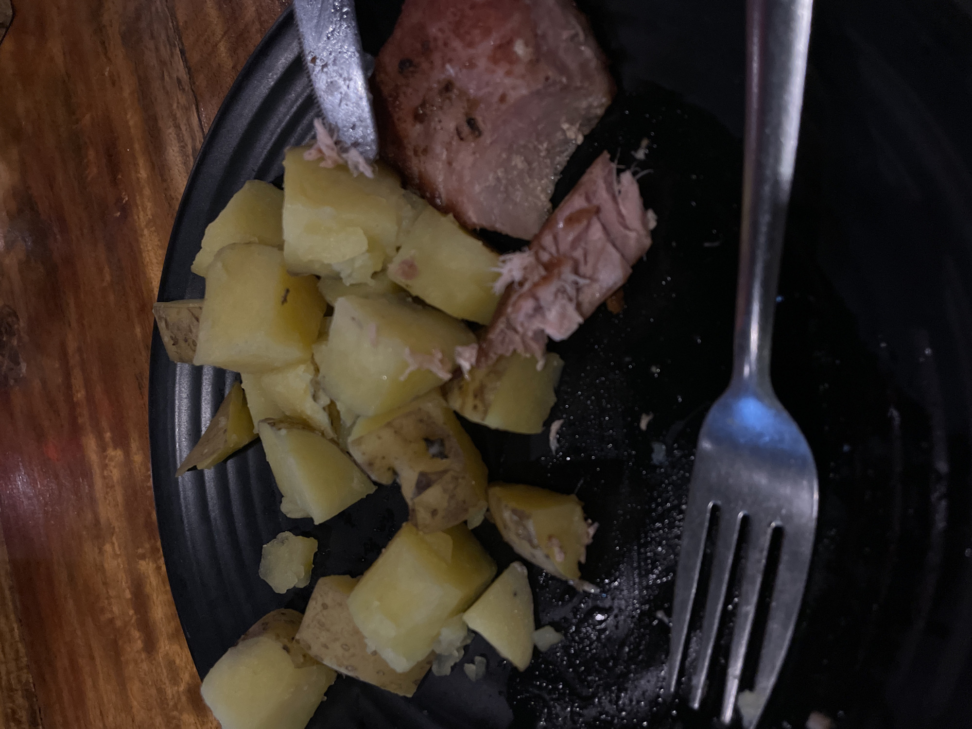 The image shows a close-up of a black plate of food on a wooden table. The plate contains several chunks of boiled or roasted potatoes, some of which appear to be cut into quarters. There is also a piece of cooked meat, possibly pork or ham, with some shredded pieces pulled away. A fork is visible in the foreground, resting on the plate, and a knife is partially visible to the right. The food appears to have been partially consumed. The lighting is dim, suggesting an indoor setting, possibly a restaurant. The background shows a wooden surface with visible grain and texture. There are no people visible in the image.