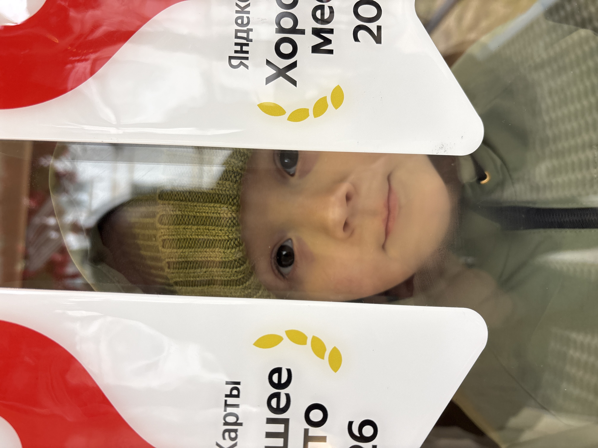 A young child in a green knit hat and light green jacket is looking through a glass partition that is split by a sign. The sign is white with a red curved shape at the top left. The words "Яндекс" (Yandex) are visible on the top sign, followed by "Хоро..." and "Ме..." with the number "20". Below, there is a yellow curved shape made of small oval shapes. The bottom sign shows "Карты" (Maps), followed by "Шее" and "то" with the number "26". The child's face is centered in the space between the signs. The scene is likely a retail environment in the city of Chaikovsky, Russia, given the presence of Cyrillic text and a sign related to Yandex, a Russian technology company.