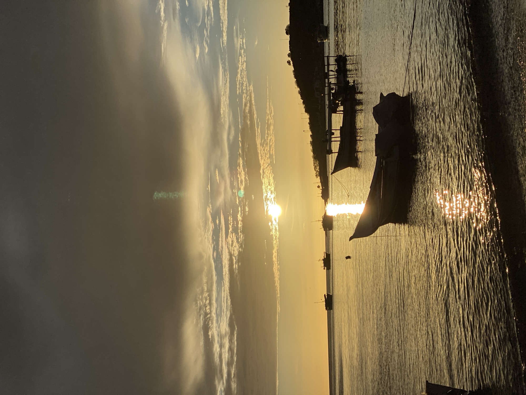 This is a vertical, medium shot of boats in the water at sunset in Juan Griego, Venezuela. The sun is low in the sky, casting a warm, golden light across the scene. The sky is filled with dramatic clouds, with some appearing dark and ominous while others are illuminated by the setting sun. The water is calm, with gentle ripples reflecting the sunlight. Several boats are visible in the water, some anchored and others appearing to be in motion. In the foreground, a small boat is tethered to a rope, with its silhouette clearly defined against the water. Further out, larger boats are seen in silhouette against the bright horizon. The background shows a hilly coastline with some structures and vegetation. The overall impression is one of a peaceful and serene evening on the water.