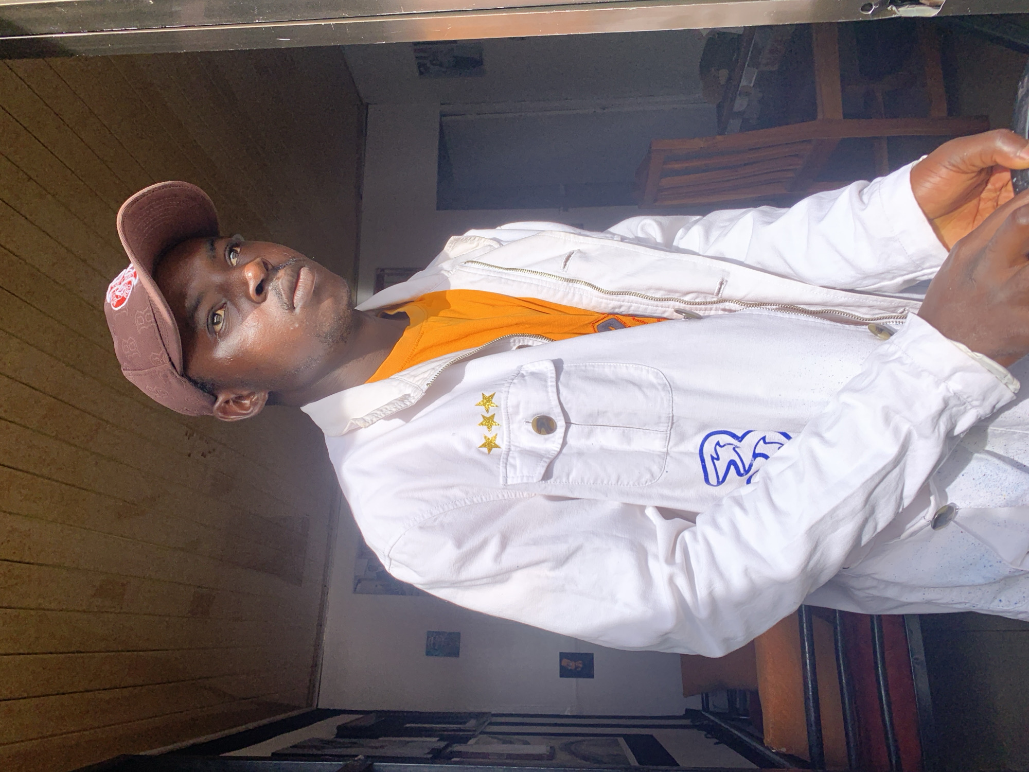 A young man with dark skin is captured in a brightly lit indoor setting, looking directly at the viewer. He wears a casual outfit consisting of a brown baseball cap, a vibrant orange t-shirt, and a white long-sleeved jacket.
His features include light-colored eyes, possibly hazel or light brown, and a subtle mustache. The brown baseball cap has a white, partially visible logo on its front. The white jacket is detailed with a zipper down the front, a chest pocket adorned with three small gold star-shaped pins, and a distinctive blue stylized logo on its right sleeve.
The background suggests an interior space, with wooden paneling visible on the left side of the frame. Strong natural light, likely from a window or doorway, illuminates the man from his left, casting clear highlights and shadows. The background is otherwise somewhat dark and blurred, revealing hints of other indoor elements like a wooden chair. The man's expression is neutral and composed, as if posing for the photograph. The scene is set in Jos, Nigeria, during daylight hours.