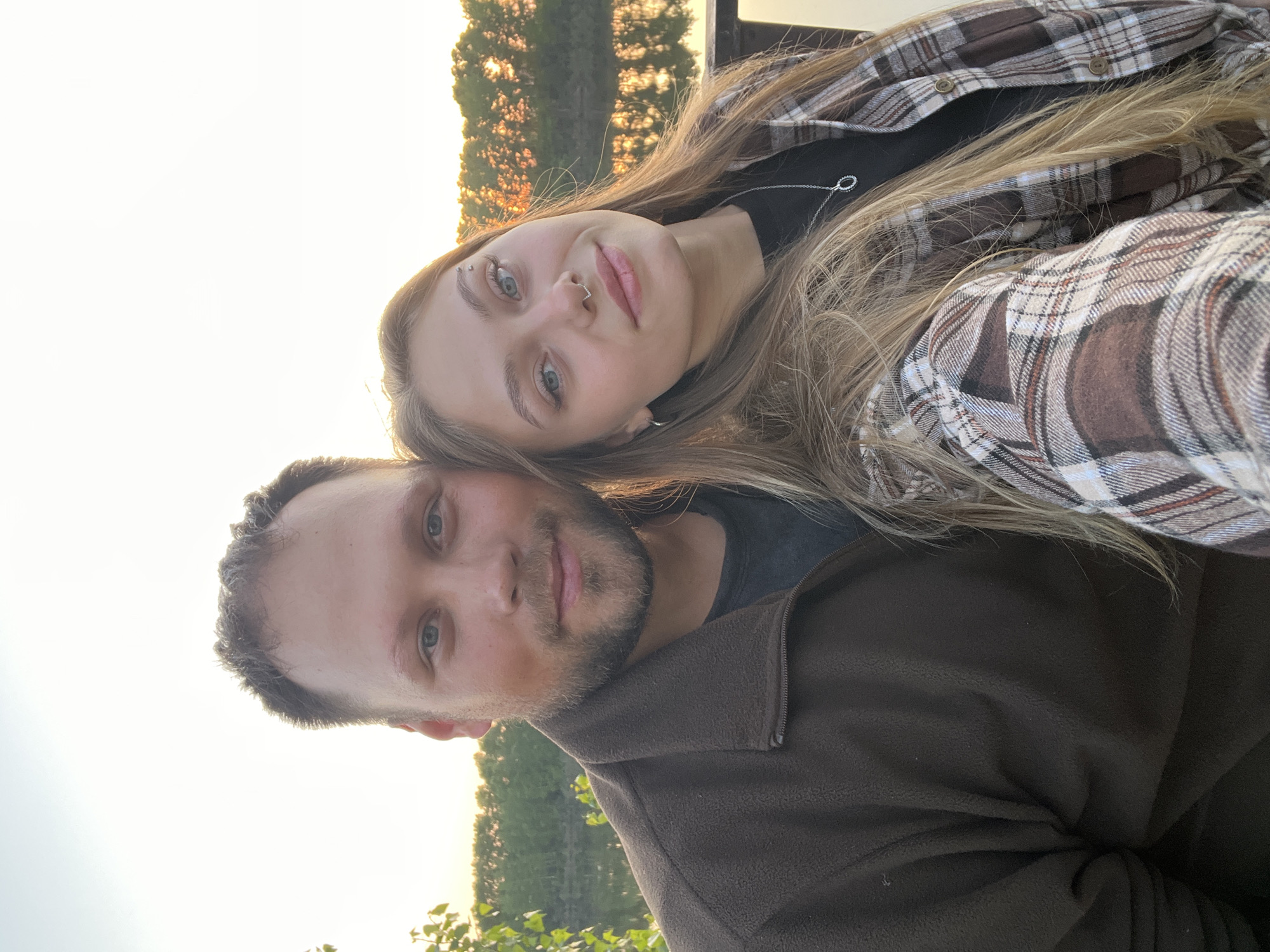 A selfie of a man and a woman. The woman has blonde hair, an eyebrow piercing, and a nose ring. She is wearing a black top and a brown and white plaid shirt. The man has short brown hair and a beard. He is wearing a brown fleece jacket. The background shows trees and a body of water. The location could be near a park in Chaikovsky, Russia.