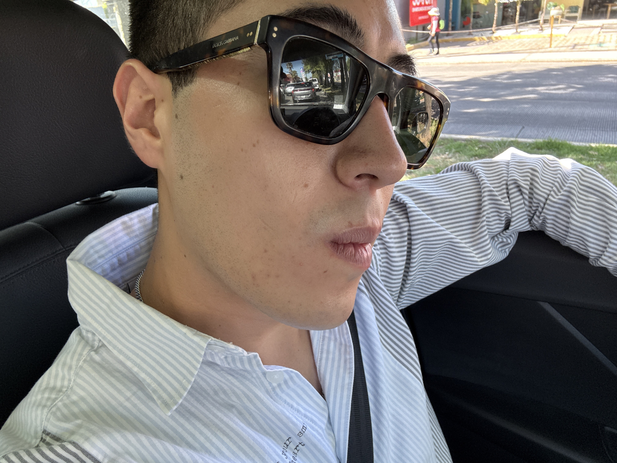 A young man is depicted in a close-up, profile shot, looking to his right, seated in what appears to be a car on a sunny day. He is wearing dark, tortoiseshell-framed sunglasses, with "DOLCE & GABBANA" visibly inscribed on the arm. His attire consists of a light blue and white striped button-down shirt. A black seatbelt is diagonally across his chest, and a silver chain necklace is partially visible around his neck.
The reflections in his sunglasses offer a glimpse into the surrounding environment: a busy street scene featuring other cars (including a light-colored sedan in front), trees lining the road, and buildings in the distance. A red sign with white text is also faintly reflected. These details suggest an urban or suburban setting, indicating the car is either driving or stopped in traffic. The man's mouth is slightly open, and his expression appears neutral, possibly observant.
The car's interior is visible, featuring a black headrest behind him and a black door panel to his right. Sunlight illuminates his face and the car's interior, suggesting clear weather conditions. On the lower front of his shirt, faint and partially obscured white text reads, "Your Smile Your Heart". The overall scene captures a moment of travel in Puebla, Mexico, during daylight hours.