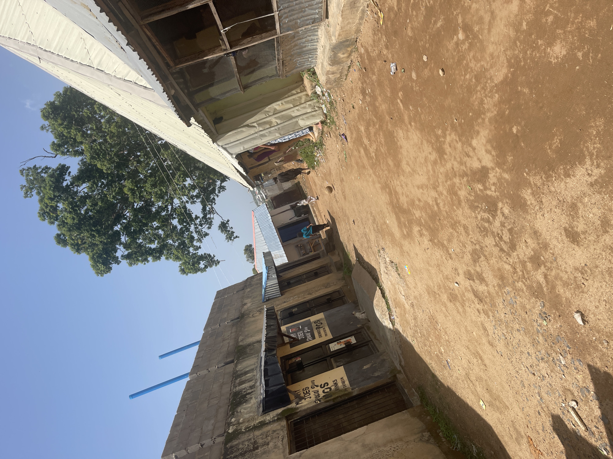 This image depicts a street scene in Jos, Nigeria, with a focus on a row of small shops. The ground is unpaved, dusty, and appears to be composed of reddish-brown soil with scattered stones and debris. The buildings lining the street are a mix of simple structures. On the left, there is a concrete building with a textured block wall, featuring several shop fronts with metal doors and awnings. Signs are visible above some of these shops, including one for "MUMMY JOES SPECIAL STORE" advertising "DRINKS PROVISION". Two blue pipes protrude from the top of the concrete wall on the left.
To the right, there are additional structures with corrugated metal roofs and walls. One building has a weathered, light-colored metal roof and a section with what appears to be a window covered in wire mesh. Another structure further down the street has a blue corrugated metal roof. Several people are present in the scene. In the foreground, a small white cat is visible near the entrance of one of the shops. Further down the street, a few individuals are walking, and a soccer ball is in the dirt. The overall impression is of a busy, but somewhat informal, commercial area. The sky above is clear blue.