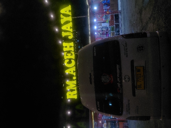 The image depicts a nighttime scene in Saree, Indonesia, featuring the rear of a white Toyota Hiace van parked in front of an illuminated establishment. The van bears an Indonesian license plate "BL 7701 NF" and numerous stickers on its rear window, including text "B. ACEH," "MEDAN," "pt. pr@m@m," a contact number "0822 6702 2006," and a vehicle inspection number "NOMOR KIR A673825000793." Above the van, a large, bright yellow neon sign spells out "RM. ACEH JAYA." To the right of the van, a series of open-air stalls are brightly lit, displaying various goods such as fruits, vegetables, and packaged items. String lights are visible across the upper portion of the scene. The ground where the van is parked appears to be a dark, possibly paved or gravel surface. No individuals are visible.