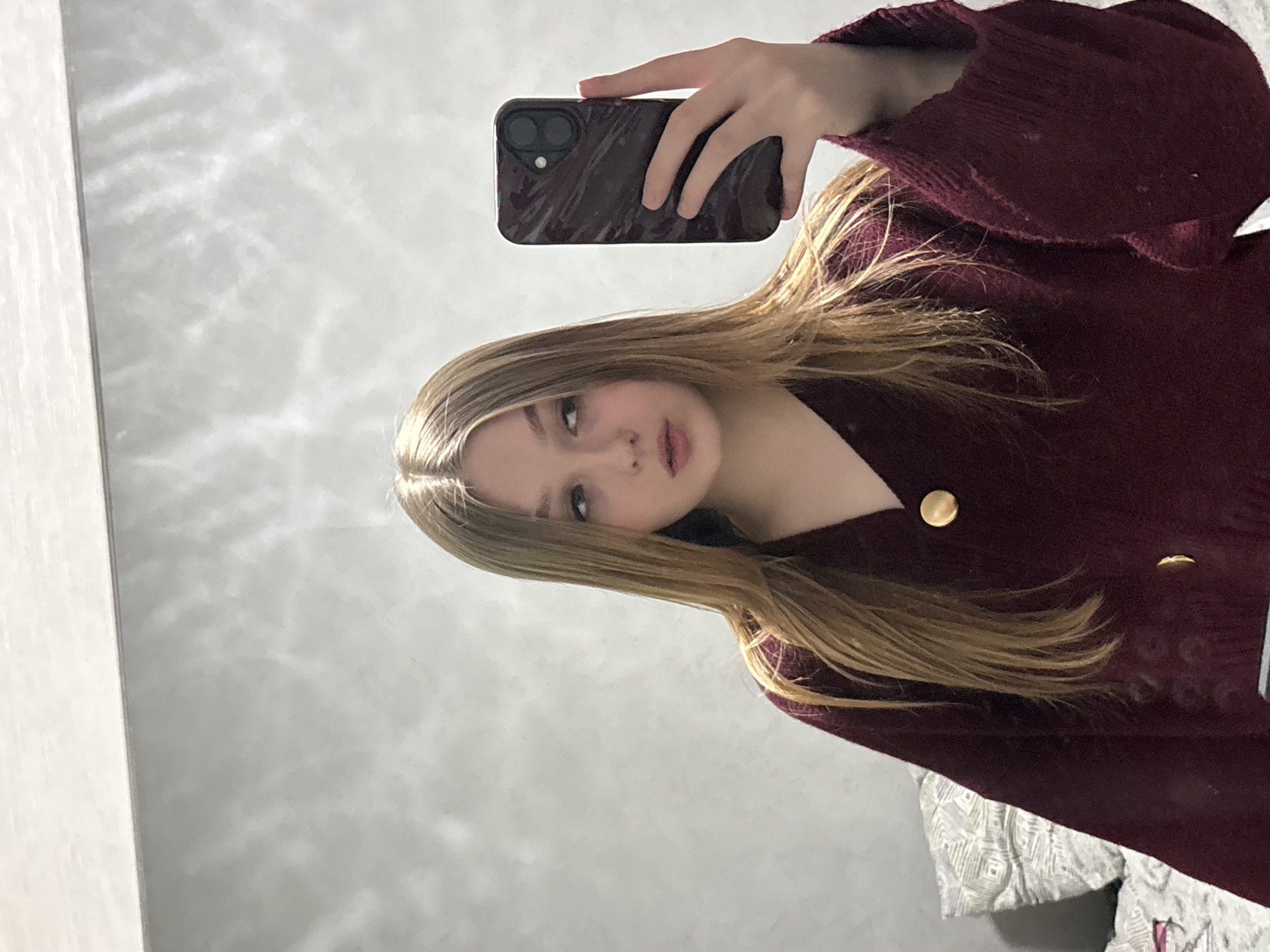 A young woman with long blonde hair is taking a selfie in a mirror. She is holding a dark-colored smartphone with a marble-patterned case. She is wearing a burgundy sweater with gold buttons. The reflection in the mirror shows a gray wall. Part of a decorative pillow or blanket can be seen in the bottom right corner of the reflection. The location in the city of Chaikovsky, Russia is not determinable from the image.