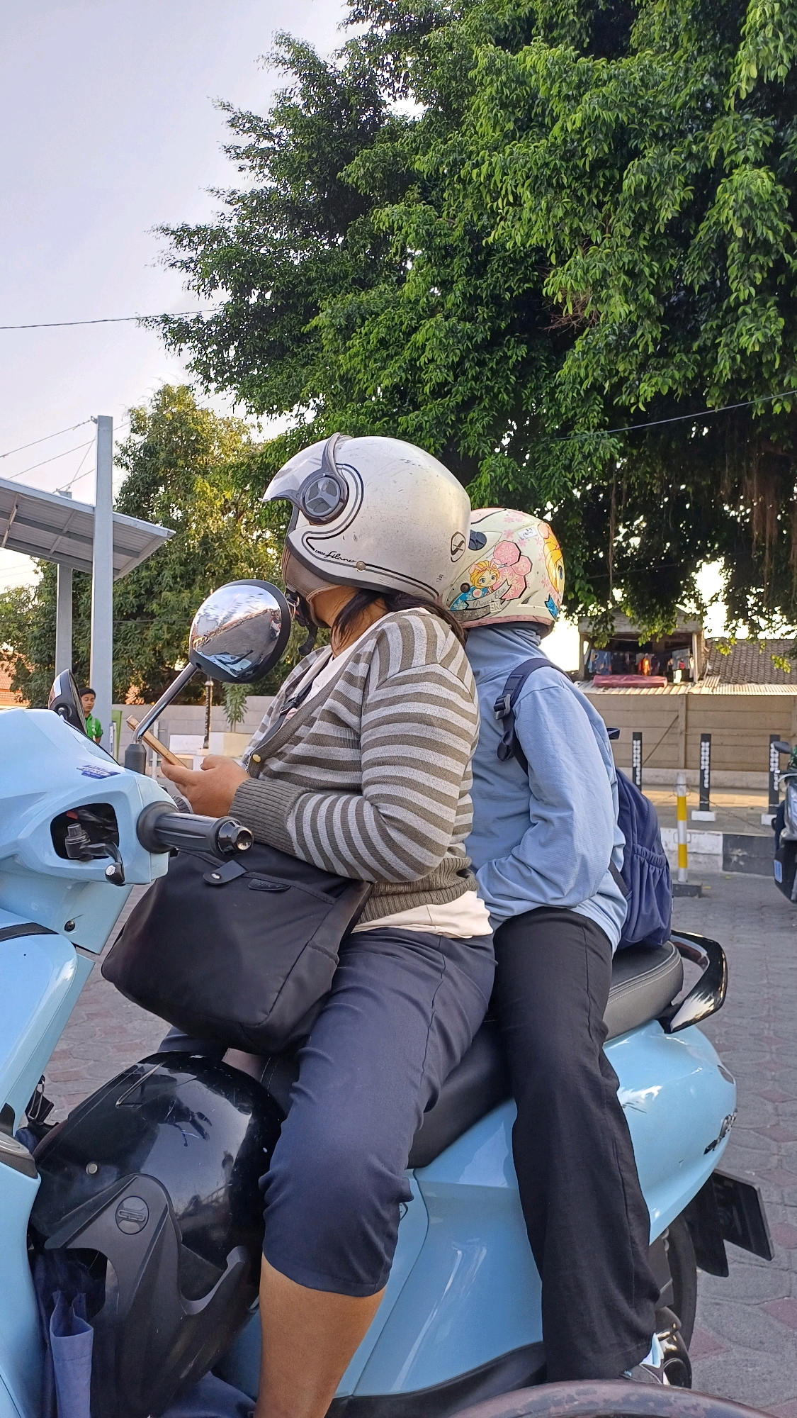 Two individuals, wearing helmets, are seated on a light blue scooter. The person in front, on the left, is looking at their phone. They are wearing a gray and white striped long-sleeved shirt and dark blue pants. A black bag is slung over their shoulder. The second person, seated behind the first, is wearing a light blue long-sleeved shirt and black pants. Their helmet has a cartoonish design featuring characters. A blue backpack is visible on their back. The scene appears to be outdoors, with lush green trees in the background. The presence of a scooter and the attire of the individuals suggest a warm climate, typical of Southeast Asia. The architectural elements in the background, such as the fence and building structures, combined with the vegetation, are consistent with a setting in Klaten, Indonesia.