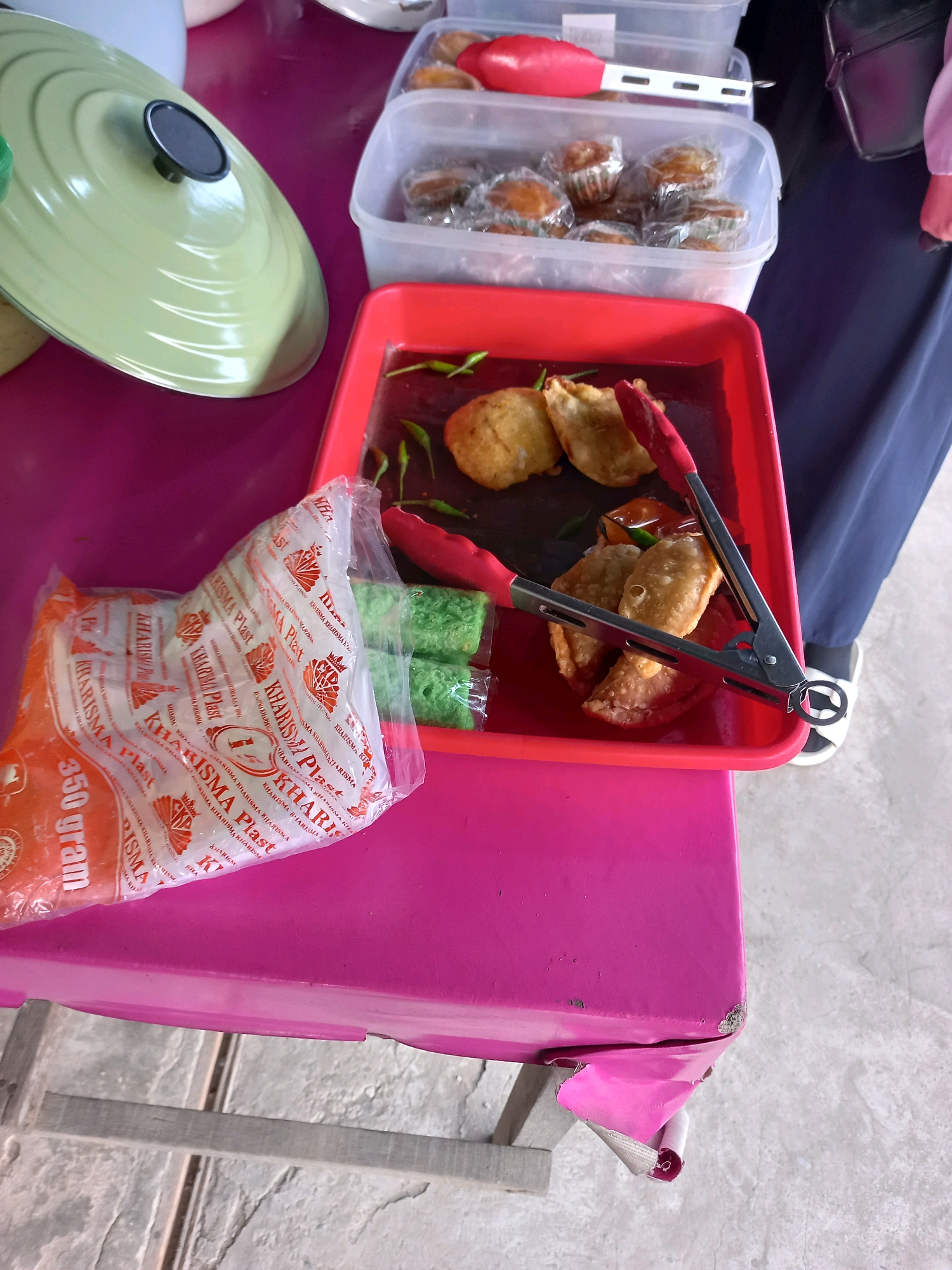 An overhead, eye-level shot shows a table laden with food items and cooking utensils, set in what appears to be an outdoor market stall in Samarinda, Indonesia. In the foreground, a bright pink table is covered with several plastic bags of snacks, one transparent bag revealing green, cube-shaped snacks wrapped in plastic, and another orange bag with text identifying it as containing 350 grams of "KHARISMA Plast". To the right of the snacks, a red plastic tray holds fried pastries, one partially obscured by red tongs. Behind this, a clear plastic container is filled with small, round pastries, possibly muffins, individually wrapped in silver foil and then plastic. A light green pot lid with a black handle rests on the left side of the table, its surface reflecting light. The background is blurred, but a person's dark blue skirt and white sandals are visible on the right. The scene is illuminated by natural light.